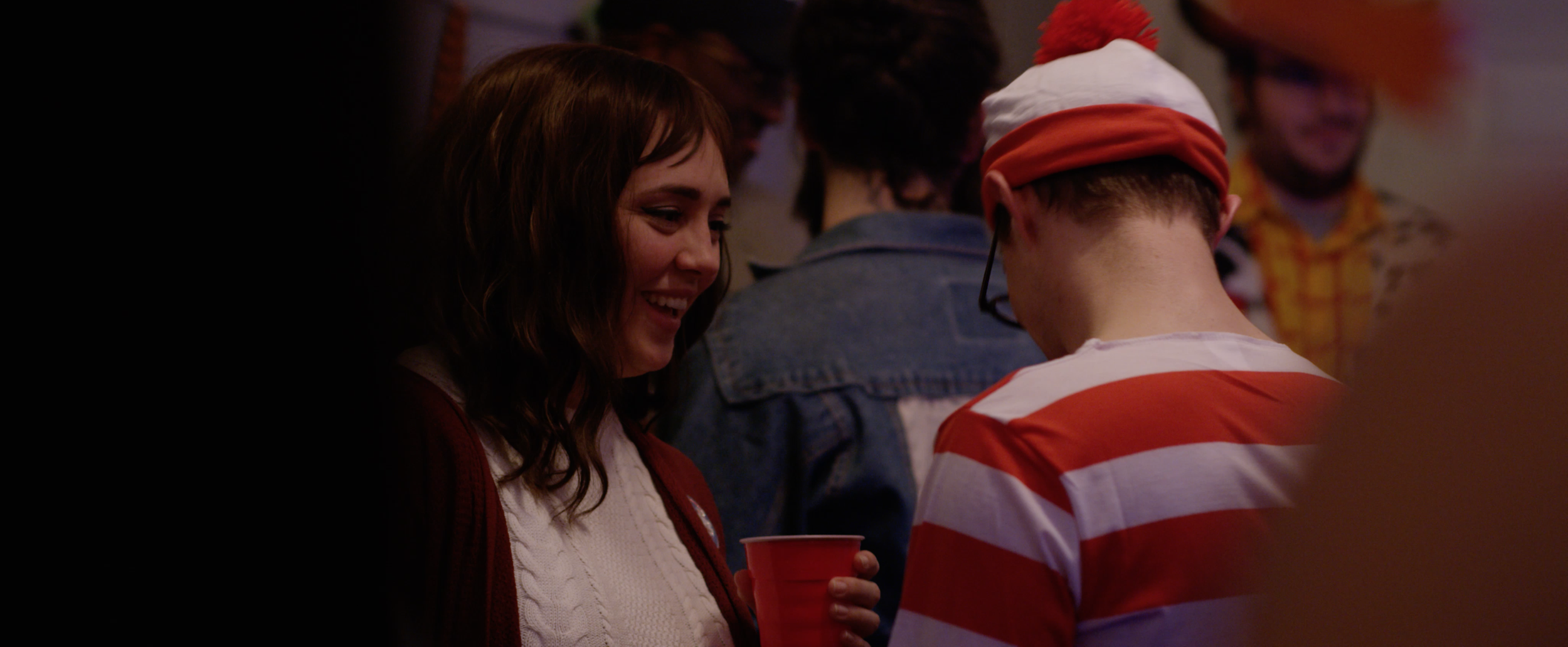 A woman and a man in a party setting chatting, with people in the background. The woman is smiling and holding a red cup, while the man is wearing glasses and a red and white striped shirt, with a white cap with a red pom-pom.