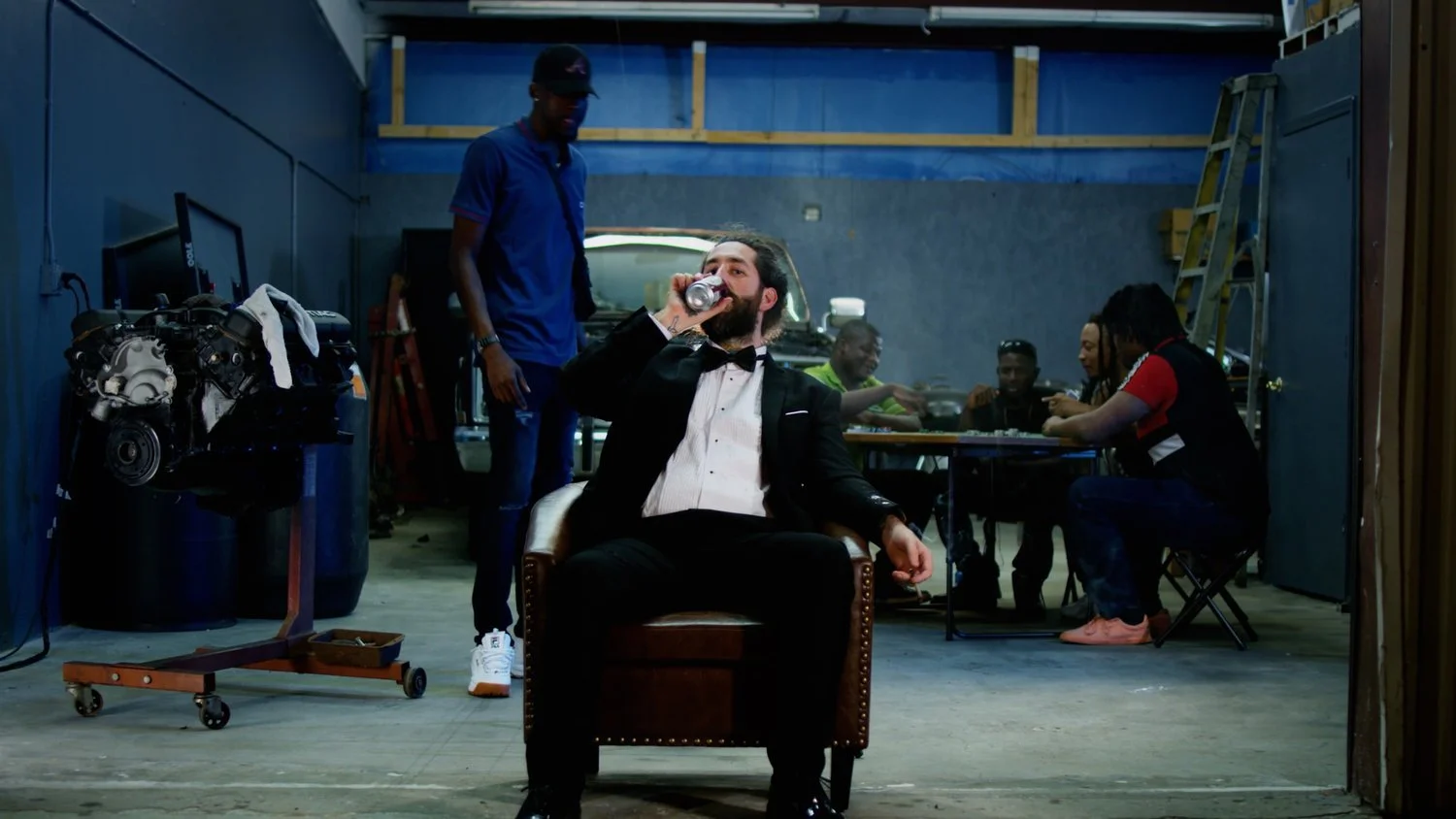 A man in a tuxedo sitting on a chair in a garage, drinking from a can, with people in the background working at a table.