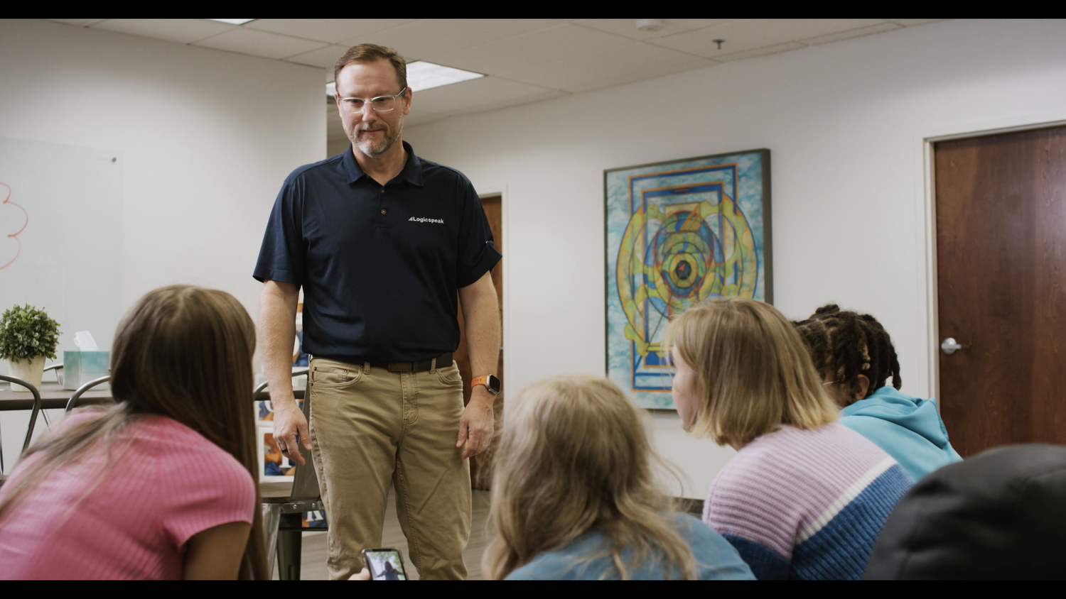 A man standing in front of a small group of children in a classroom setting. The man is wearing glasses, a navy polo shirt with a logo, beige pants, and a smartwatch. The children are sitting and listening, with one holding a phone. The classroom has