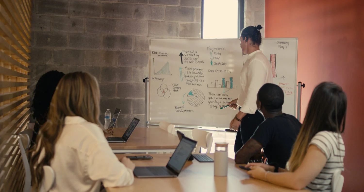 A woman presents data on a whiteboard to a group of four people seated at a table with laptops and phones in a modern conference room.
