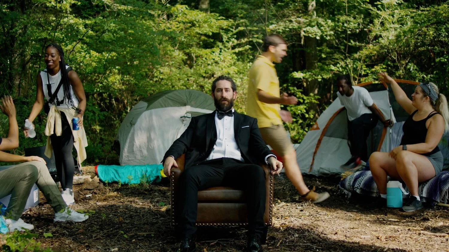 A man in a tuxedo sitting on an armchair in a woodland camping site, surrounded by tents and people socializing.