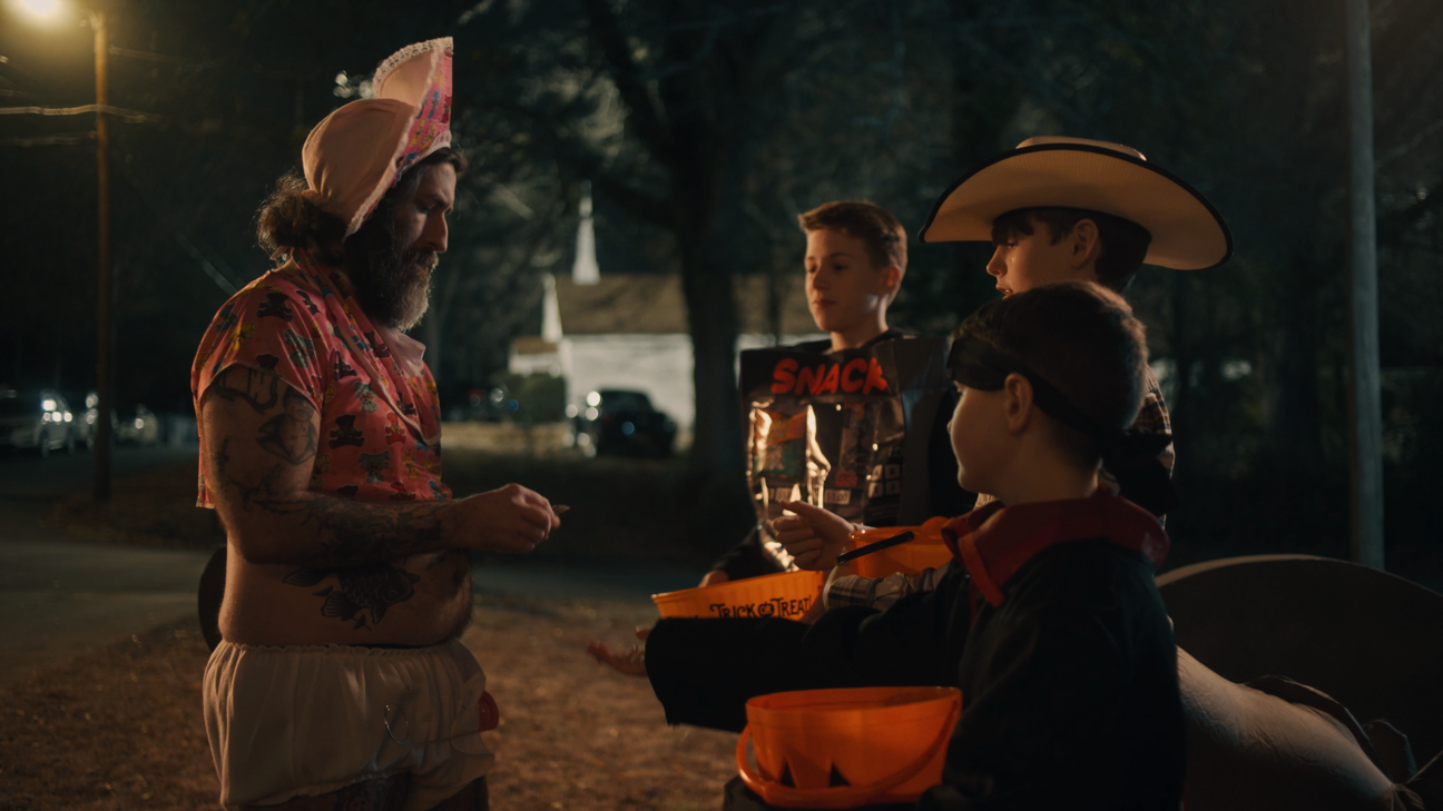 A man with tattoos wearing a colorful shirt, hat, and shorts is talking to three children wearing costumes. One child wears a cowboy hat, another a mask, and the third a bandana. They are outdoors at night with trees and a church in the background.