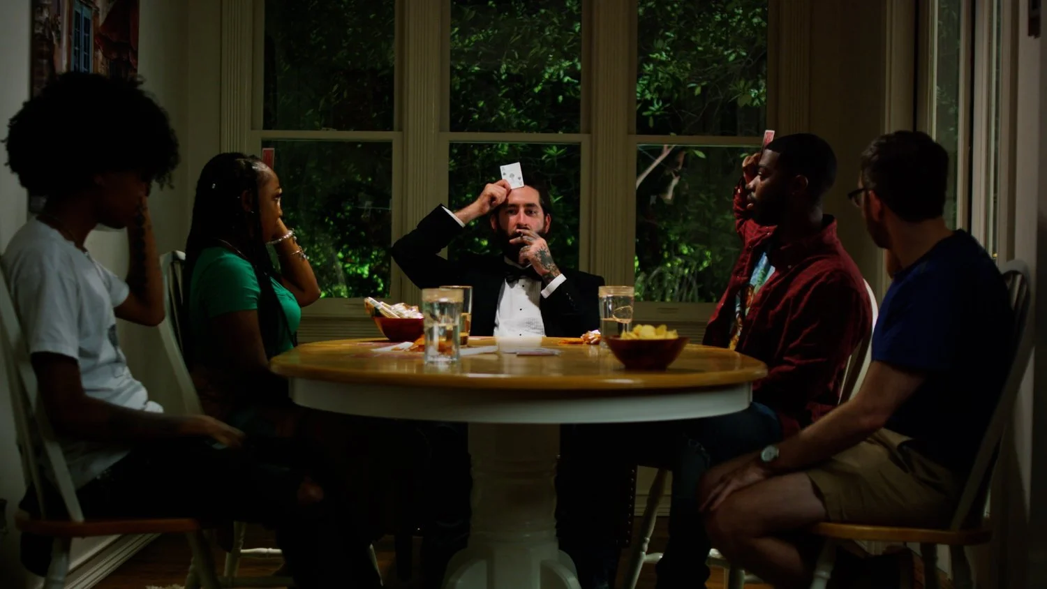 People gathered around a dining table, playing a game with cards, with food and drinks on the table, large windows showing greenery outside.