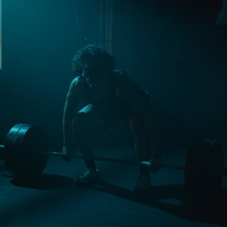 A person with curly hair preparing to lift a heavy barbell in a dimly lit gym, shadowed and focused.