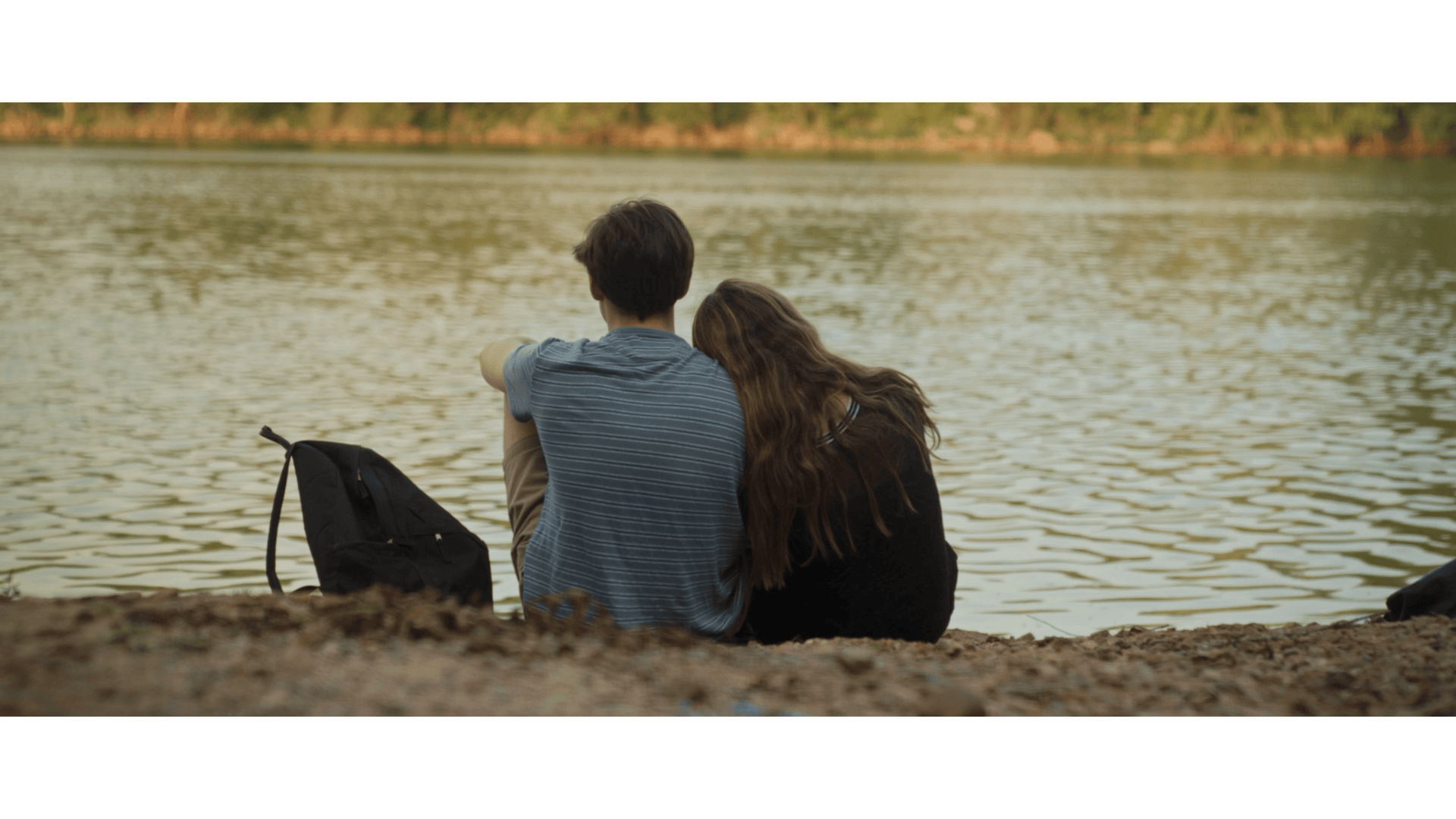 A young couple sitting on a riverbank, with the man pointing towards the water and the woman resting her head on his shoulder, a black backpack beside them.