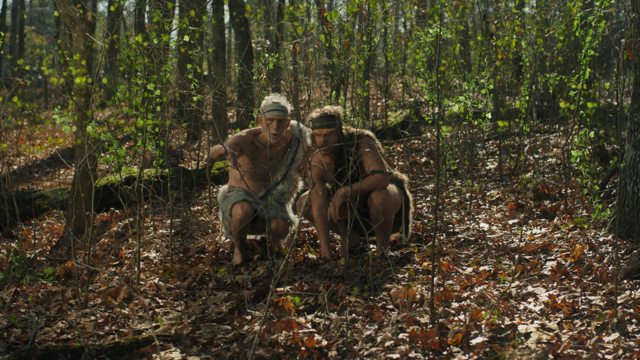 Two people dressed in primitive clothing crouch in a wooded forest, appearing to examine something on the ground.