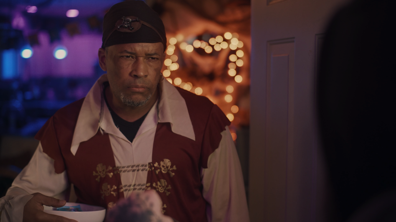 A man dressed as a pirate with a bandana and skull-printed vest looks serious, holding a bowl, in a dimly lit setting with colorful bokeh lights in the background.