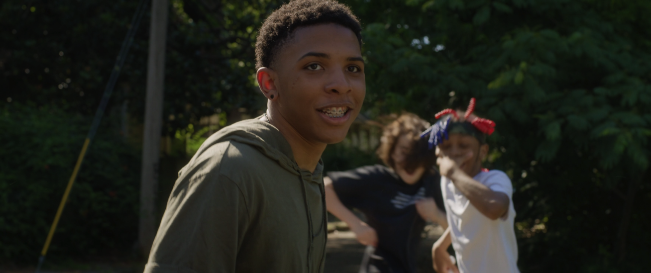 A young man with braces smiling at the camera outdoors, with two children in the background playing and laughing.