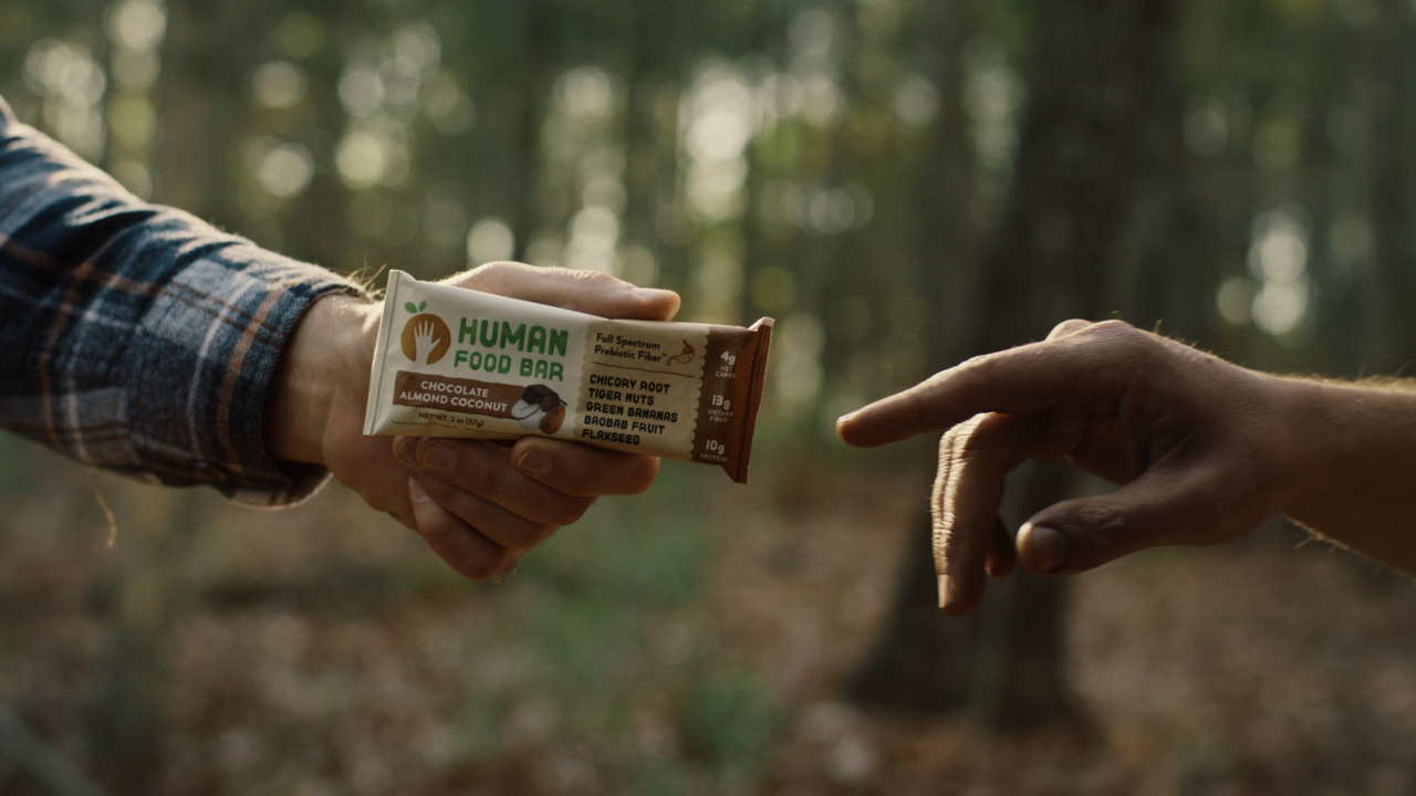 Two hands, one holding a Human Food Bar and the other reaching to take it, in a forest setting.