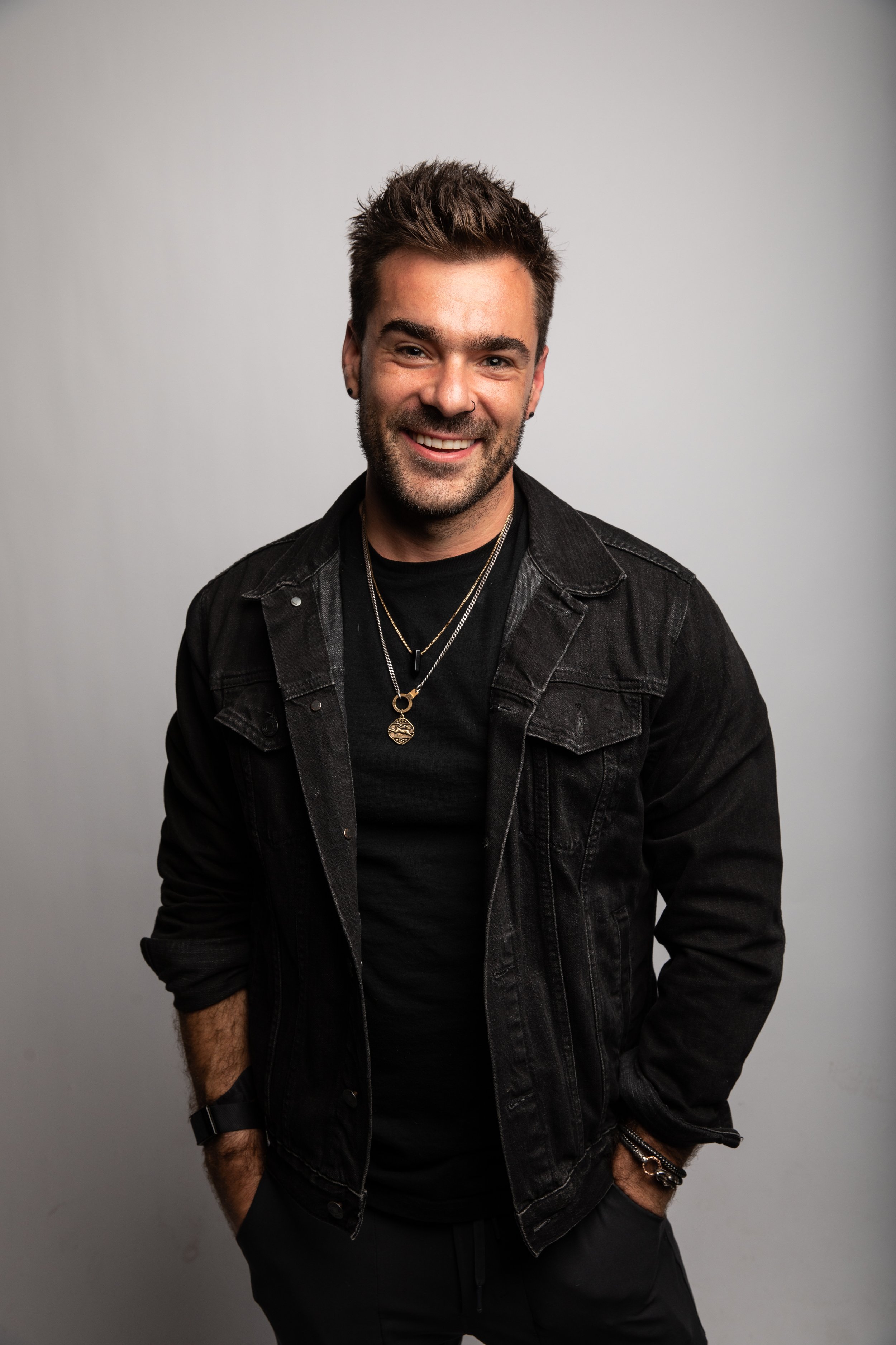 A smiling young man with dark hair and a beard, wearing a black jacket, layered necklaces, and casual black clothes, standing against a plain light grey background.