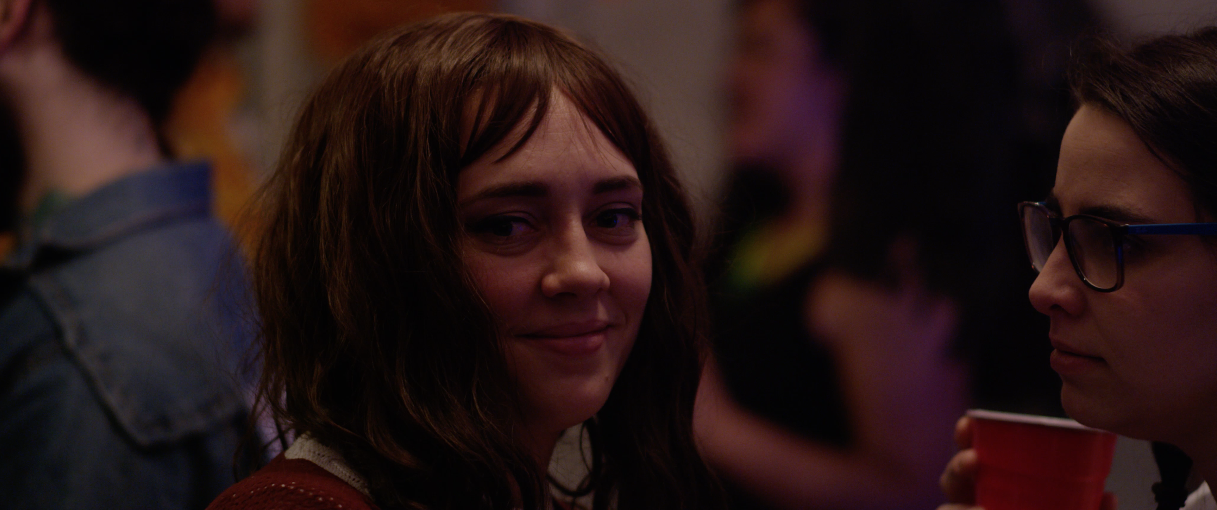 A woman with shoulder-length wavy brown hair and a subtle smile is looking towards the camera. She is in a social setting with blurred people and colorful lighting in the background. Another woman with glasses and a red cup is visible on the right si