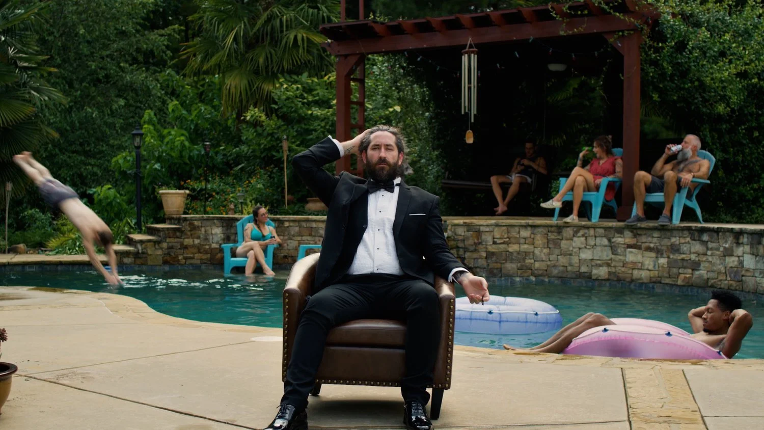 A man in a tuxedo sitting on a chair in the pool area, with one hand on his head, surrounded by people swimming, lounging, and sitting on chairs in a backyard pool setting.