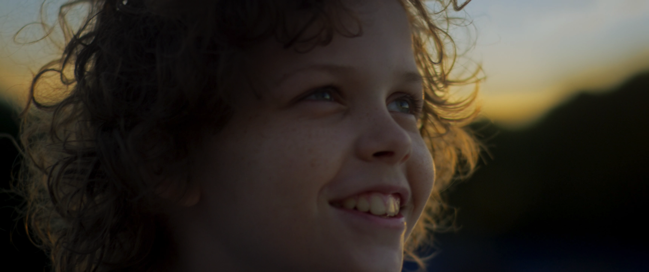 Close-up of a young boy with curly hair smiling outdoors at sunset.