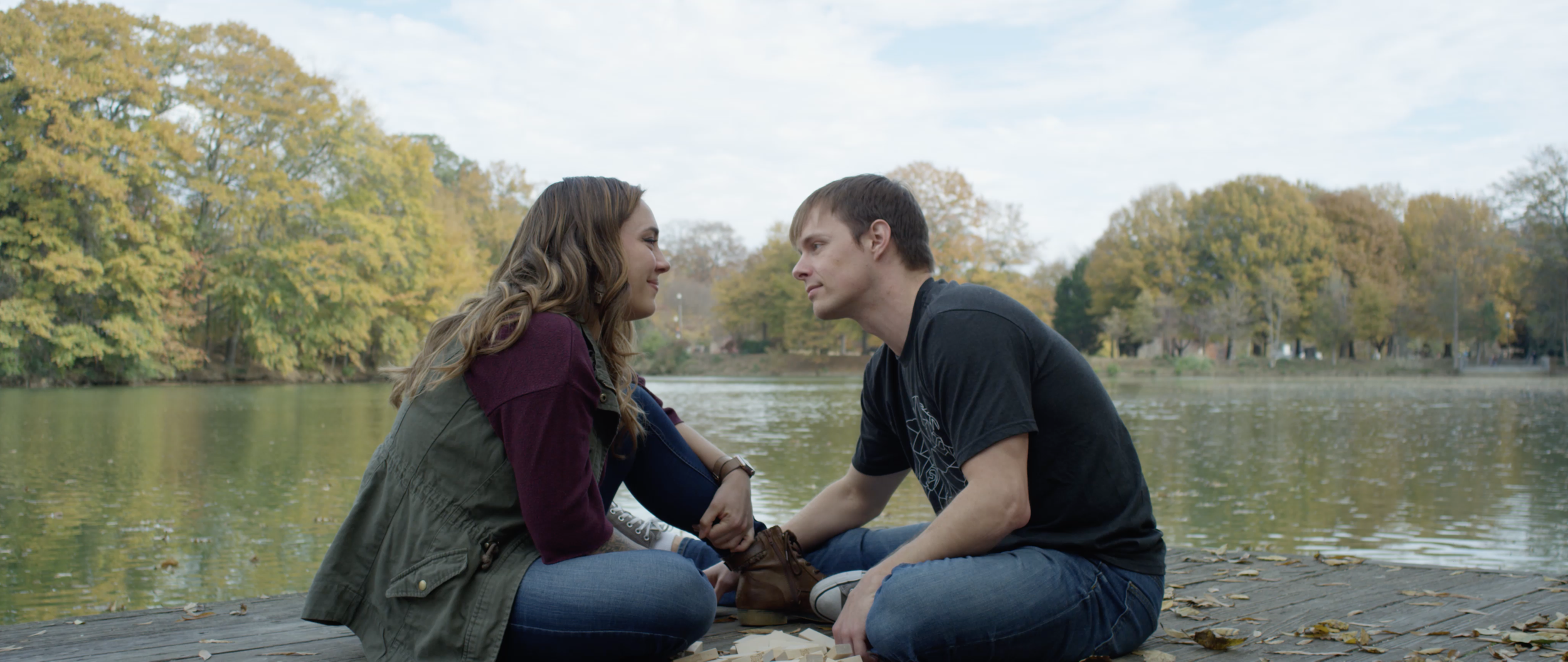 A young couple sitting on a wooden dock by a lake, facing each other closely, holding hands, surrounded by green and yellow trees during autumn.