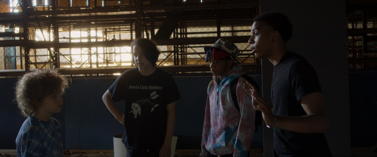 Four teenagers having a conversation indoors near a window with sunlight streaming in, in an industrial or warehouse setting.