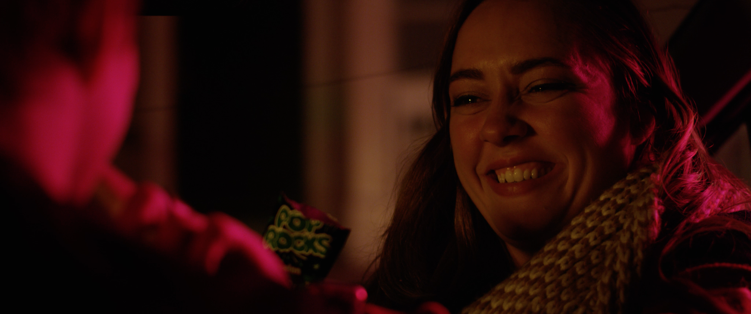 A woman with a smiling face, wearing a multicolored scarf, in a dimly lit setting with pink and reddish lighting.