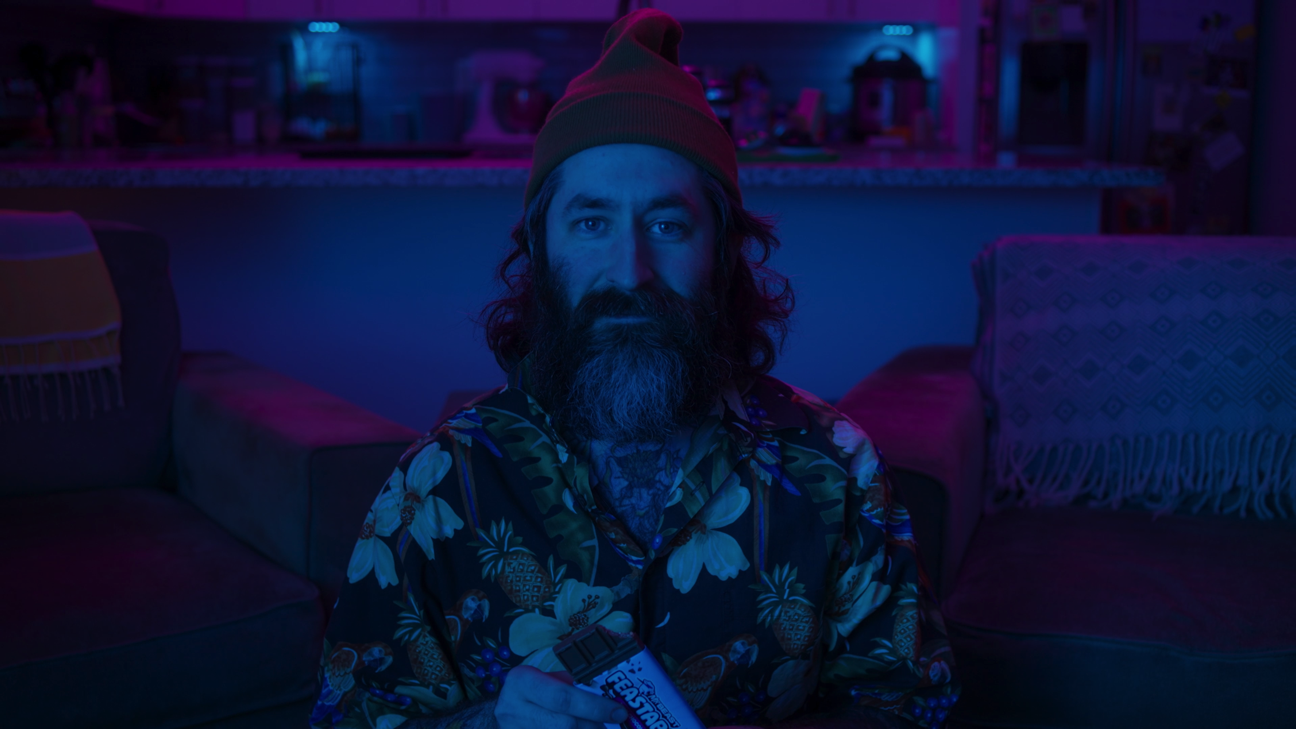 Bearded man with long hair wearing a red beanie and a colorful Hawaiian shirt sitting on a couch in a dimly lit room with purple and blue lighting, holding a box of snacks.