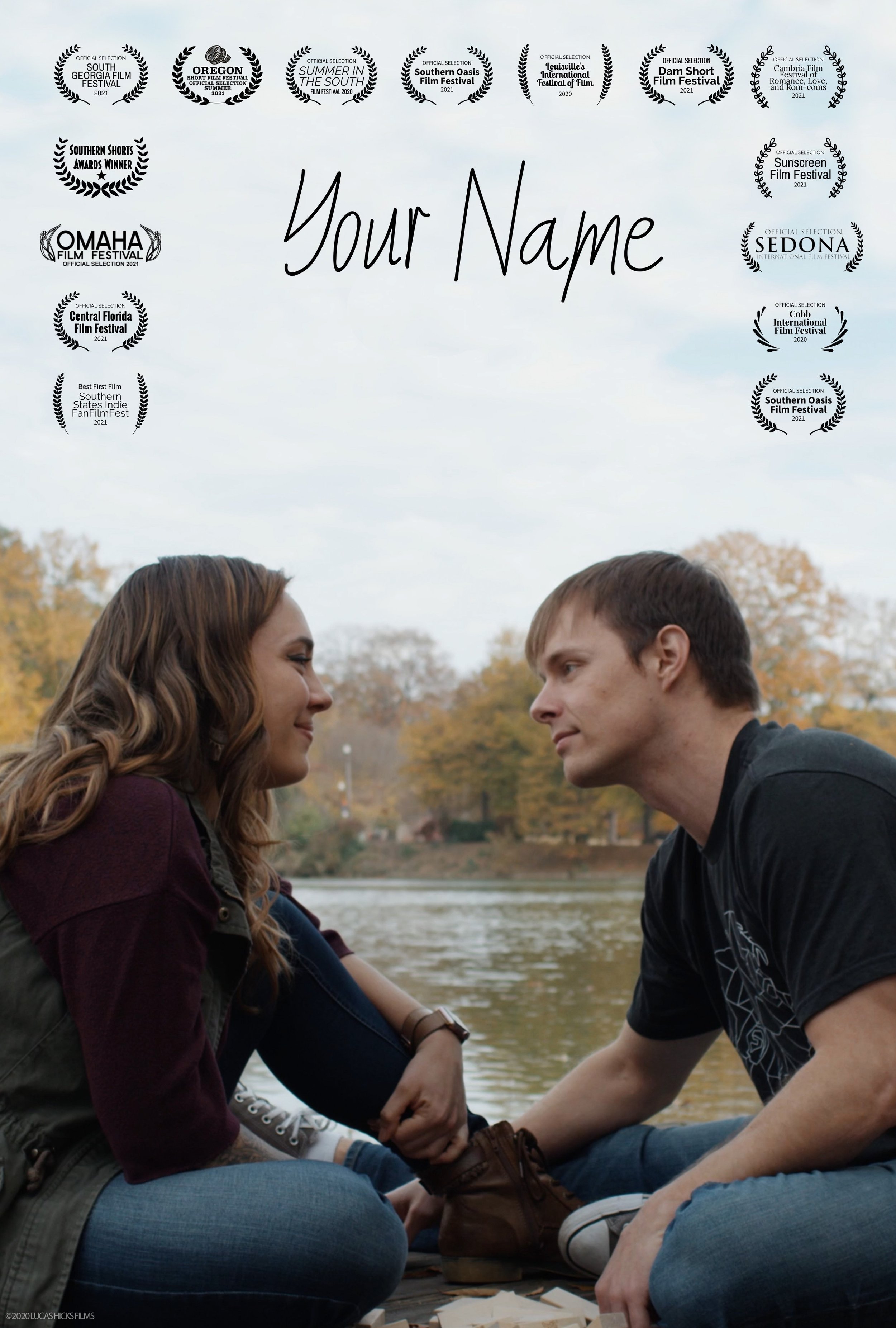 A man and woman facing each other outdoors by a lake, smiling and holding hands, with autumn trees in the background.