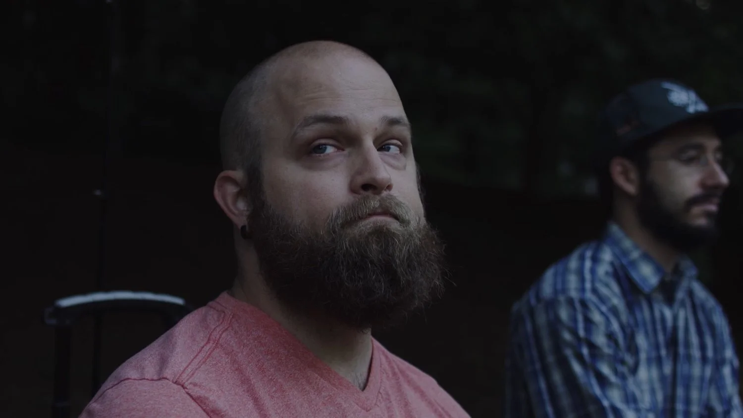 Two men sitting outdoors, one with a beard and wearing a red shirt looking towards the camera, the other with glasses and a helmet in a blue plaid shirt looking away.