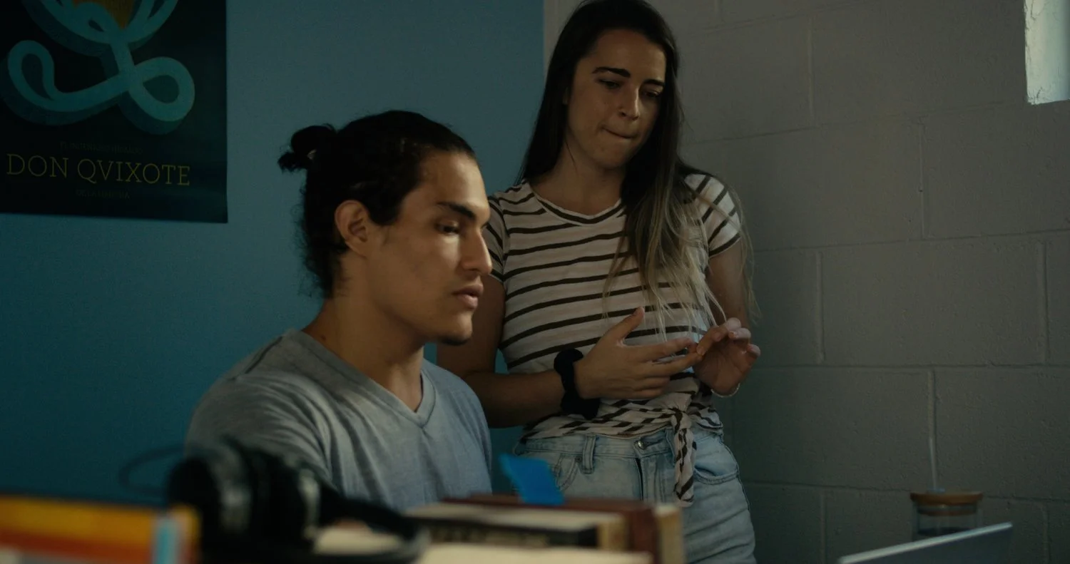 A young man sitting at a desk looking down, with a young woman standing behind him looking at her phone.