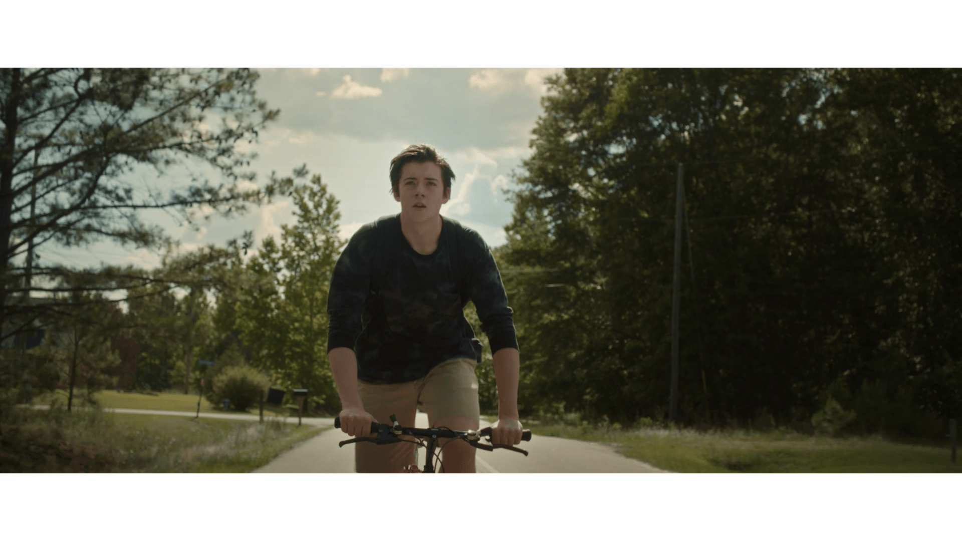 A teenage boy riding a bicycle on a paved path through a park, surrounded by trees on a sunny day.