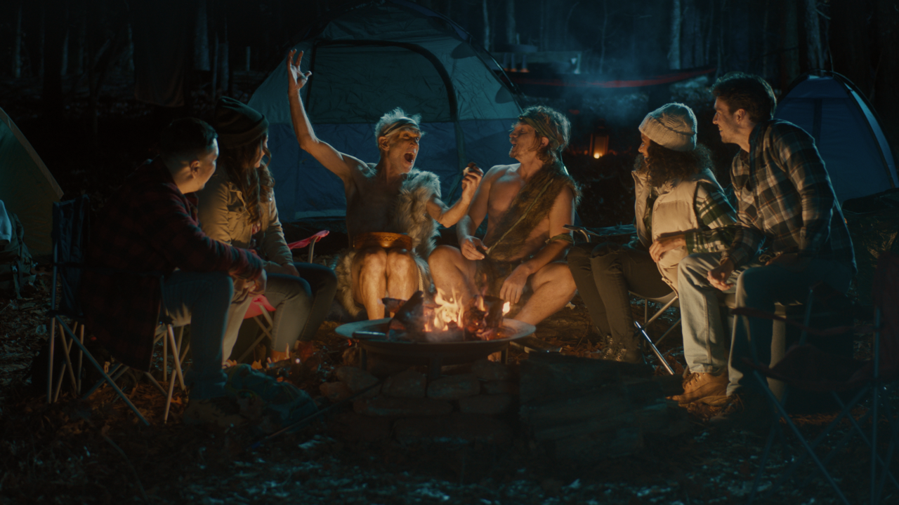 Group of people sitting around a campfire at night in a wooded area with tents in the background.