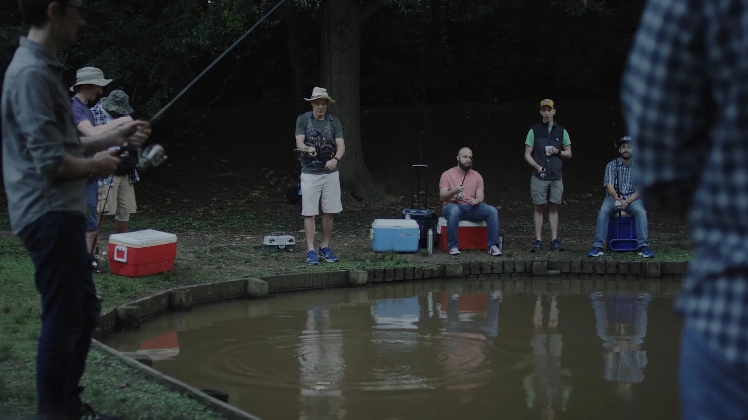 Group of people fishing by a small pond during daytime, some sitting on coolers, others standing, with fishing gear and coolers around.