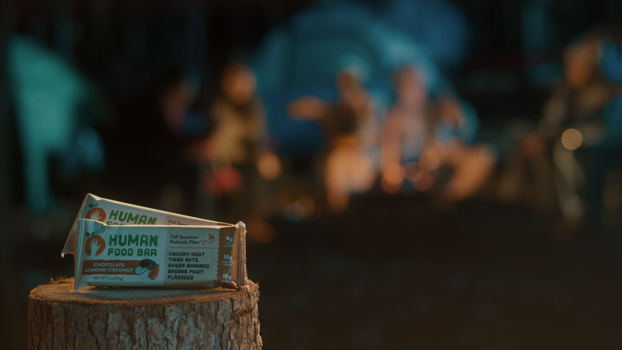 A close-up of a wrapped human food bar on a tree stump with a blurred campfire and people in the background.
