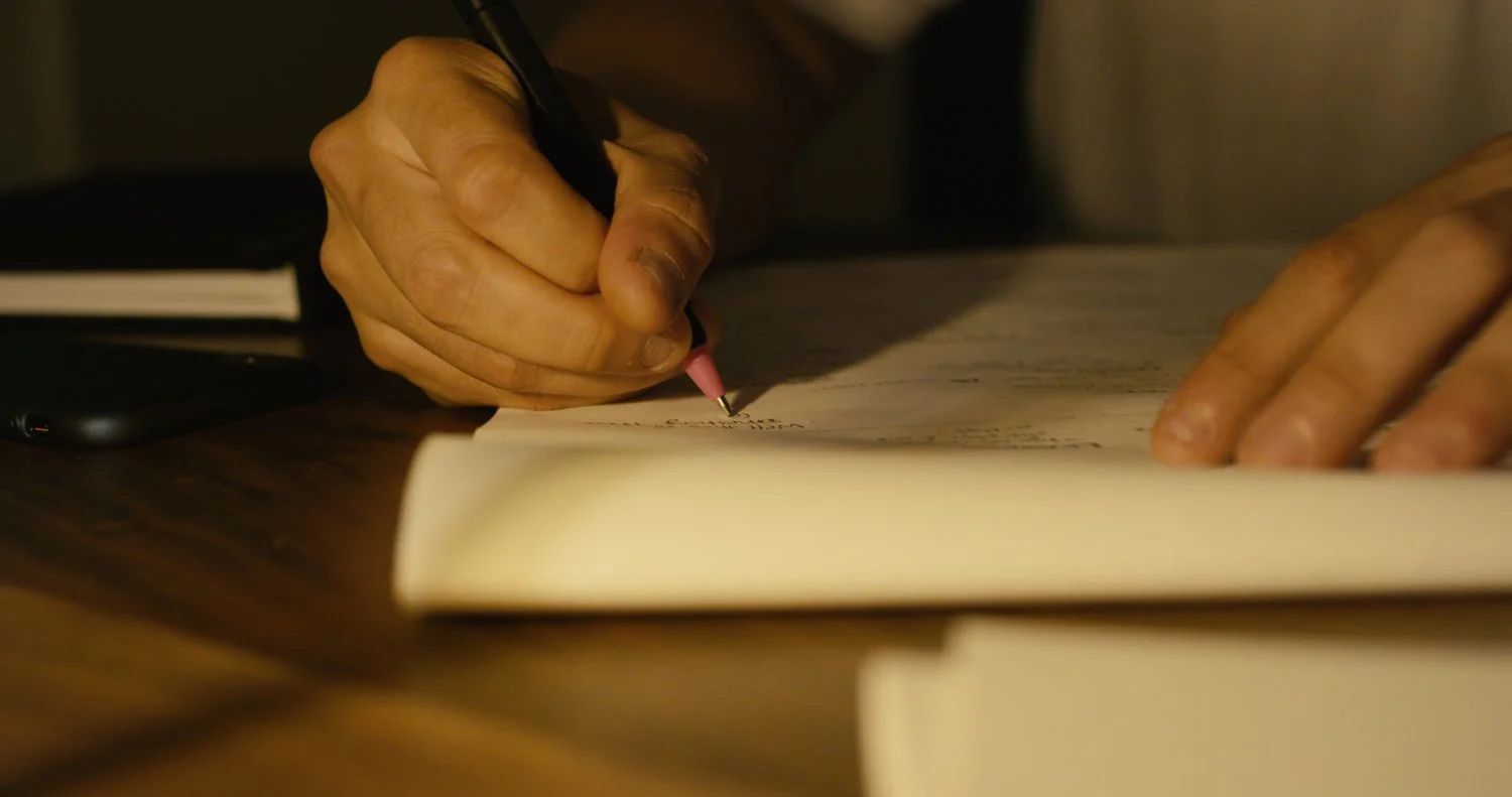 Close-up of a person writing on a piece of paper with a black pen on a wooden desk.