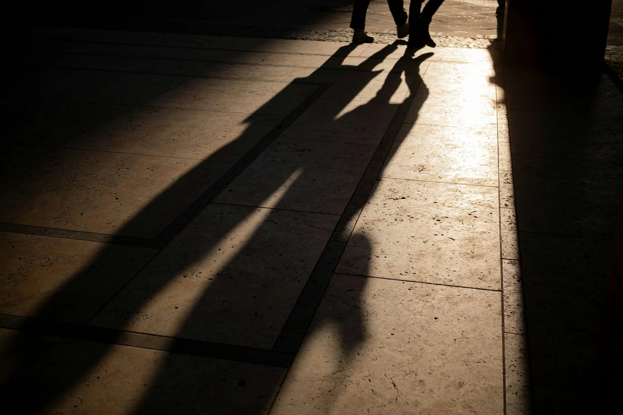 Shadows of two people walking on a sidewalk with sunlight reflecting off the pavement.