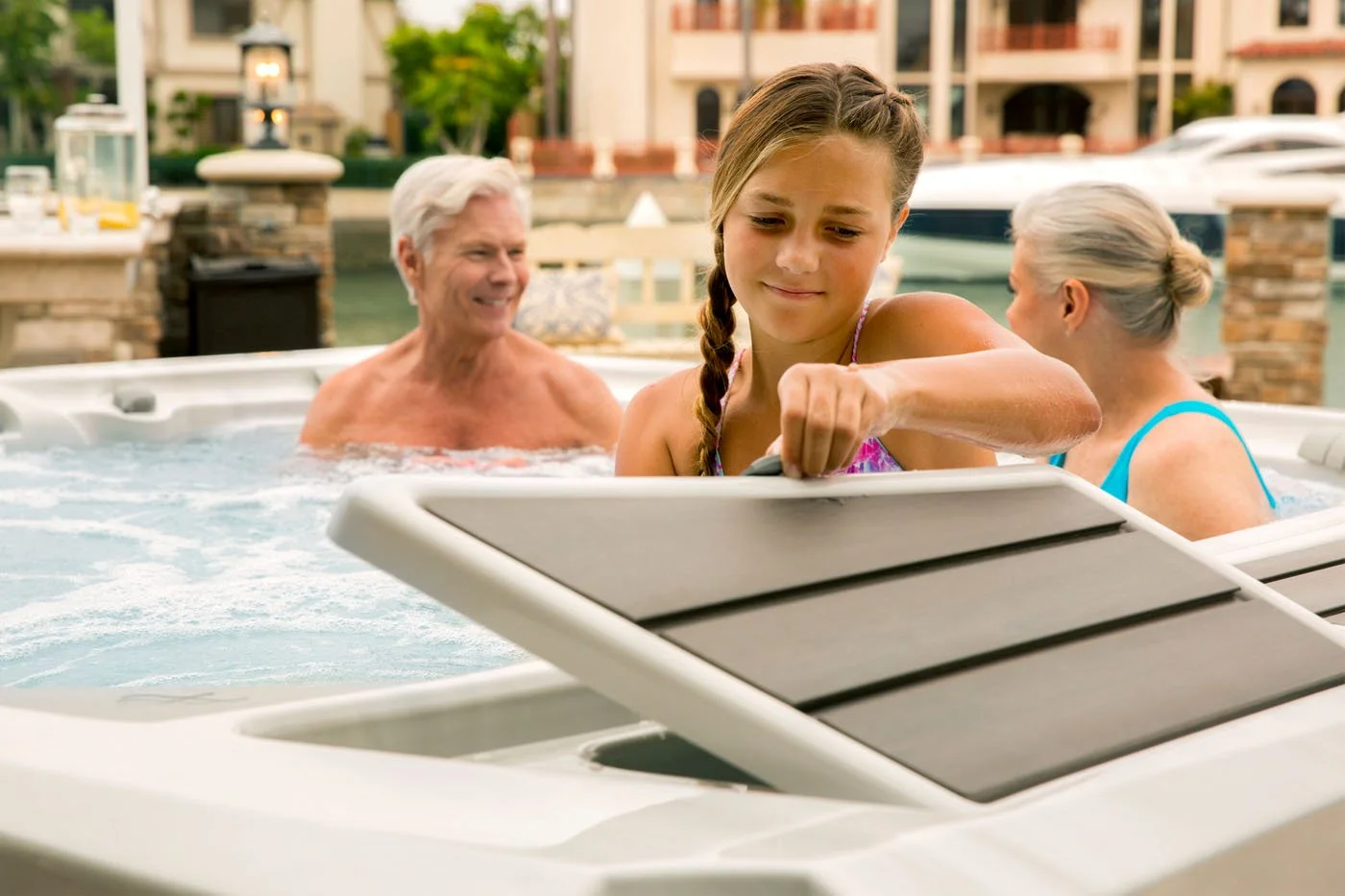 A girl opens a hot tub compartment while two older adults relax in the bubbling water outdoors.