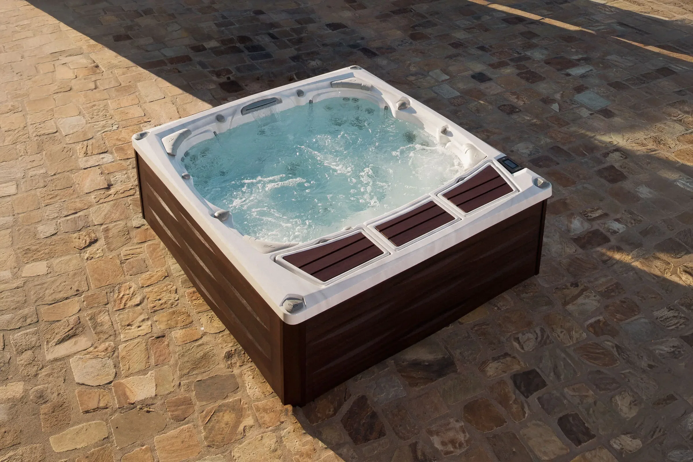 A Sundance 980 Series hot tub with bubbling water sits on a stone patio under partial shade.