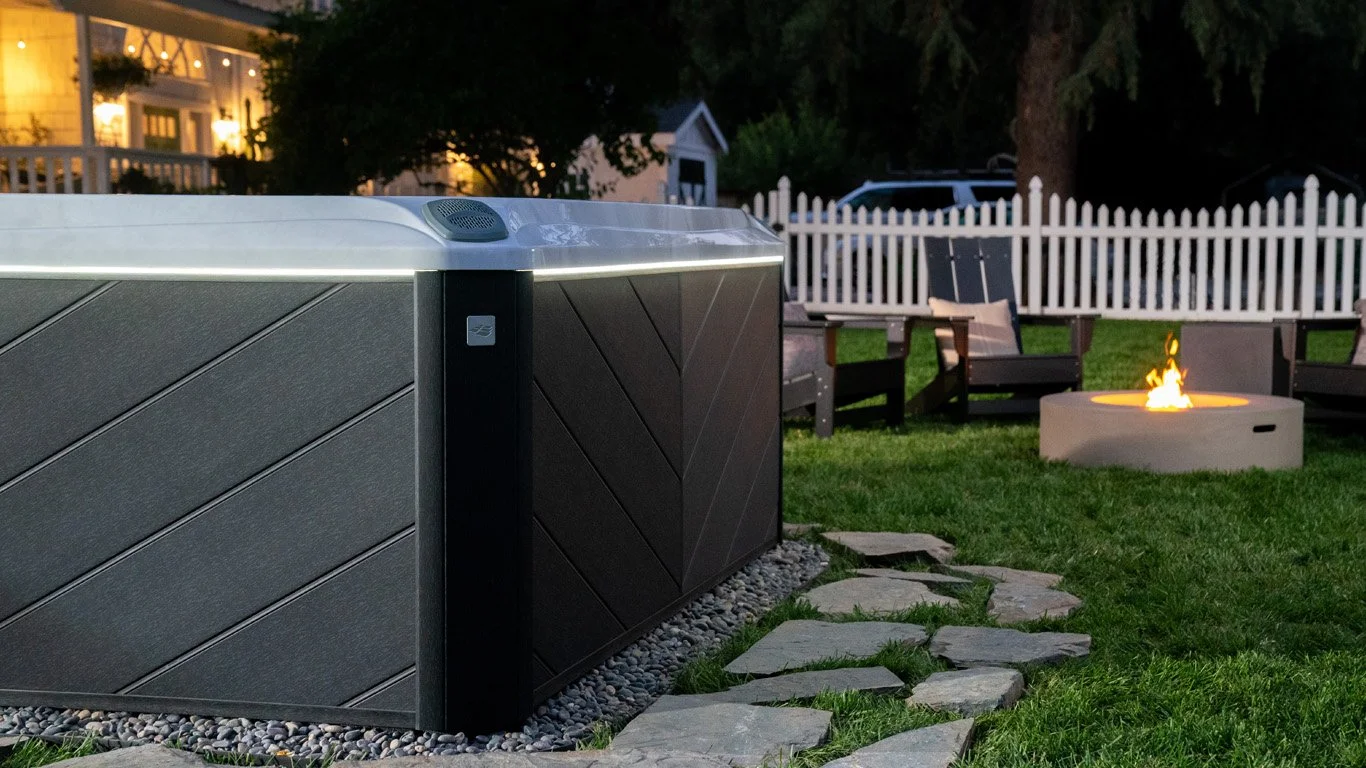 Modern hot tub with LED lights, 880 Exterior Lighting, stone path, fire pit, Adirondack chairs, and picket fence at dusk.