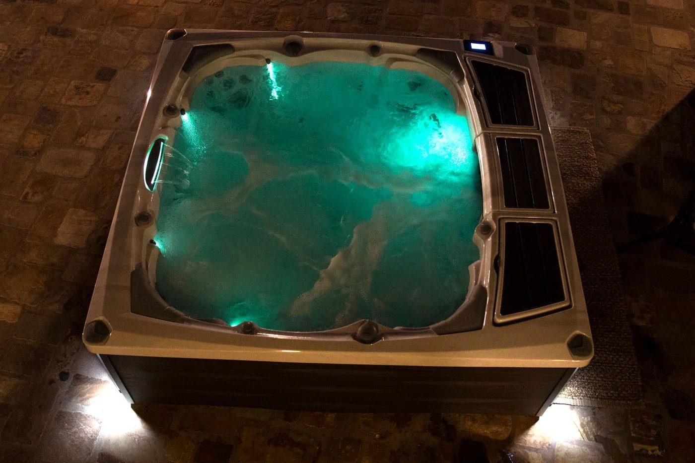 A bubbling hot tub with glowing turquoise lights, viewed from above on a stone-tiled floor at night.