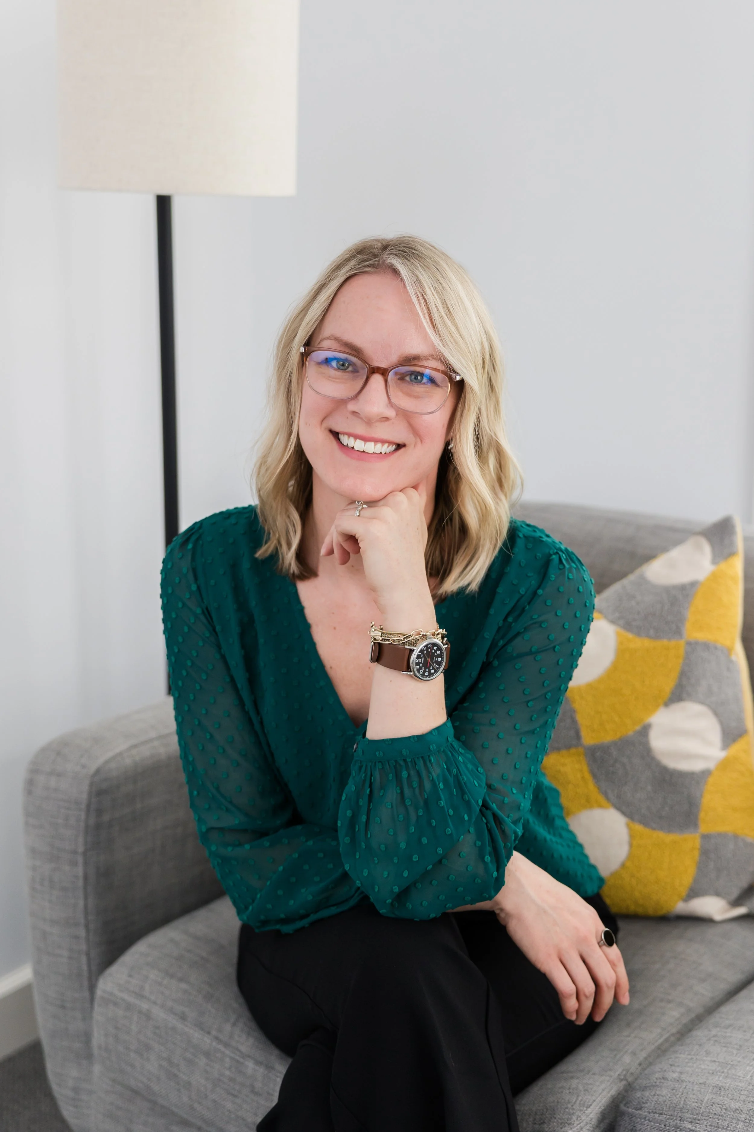 A smiling woman with blonde hair, wearing glasses, a green long-sleeve shirt, a watch, and a ring, sitting on a gray sofa with a patterned pillow in the background.