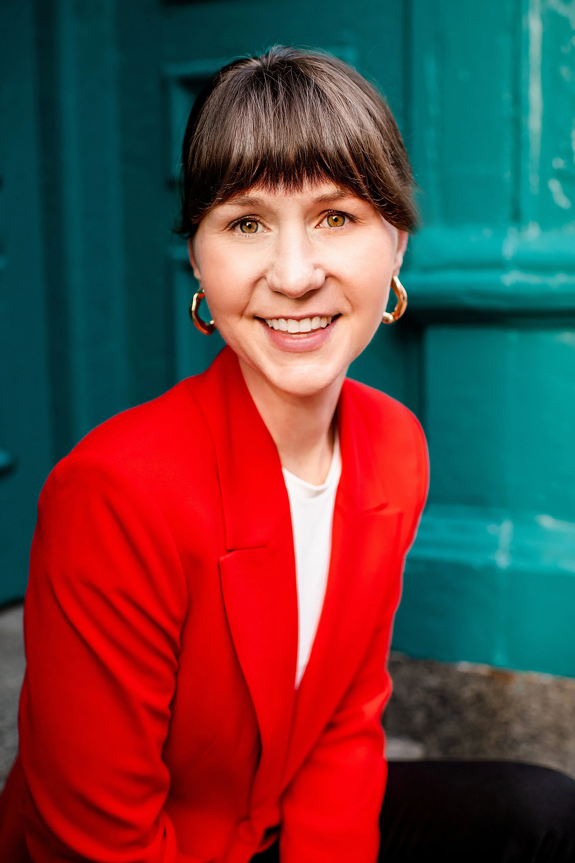 A woman with short brown hair and light brown eyes smiling at the camera, wearing a red blazer and silver hoop earrings, sitting outdoors against a greenish wall.