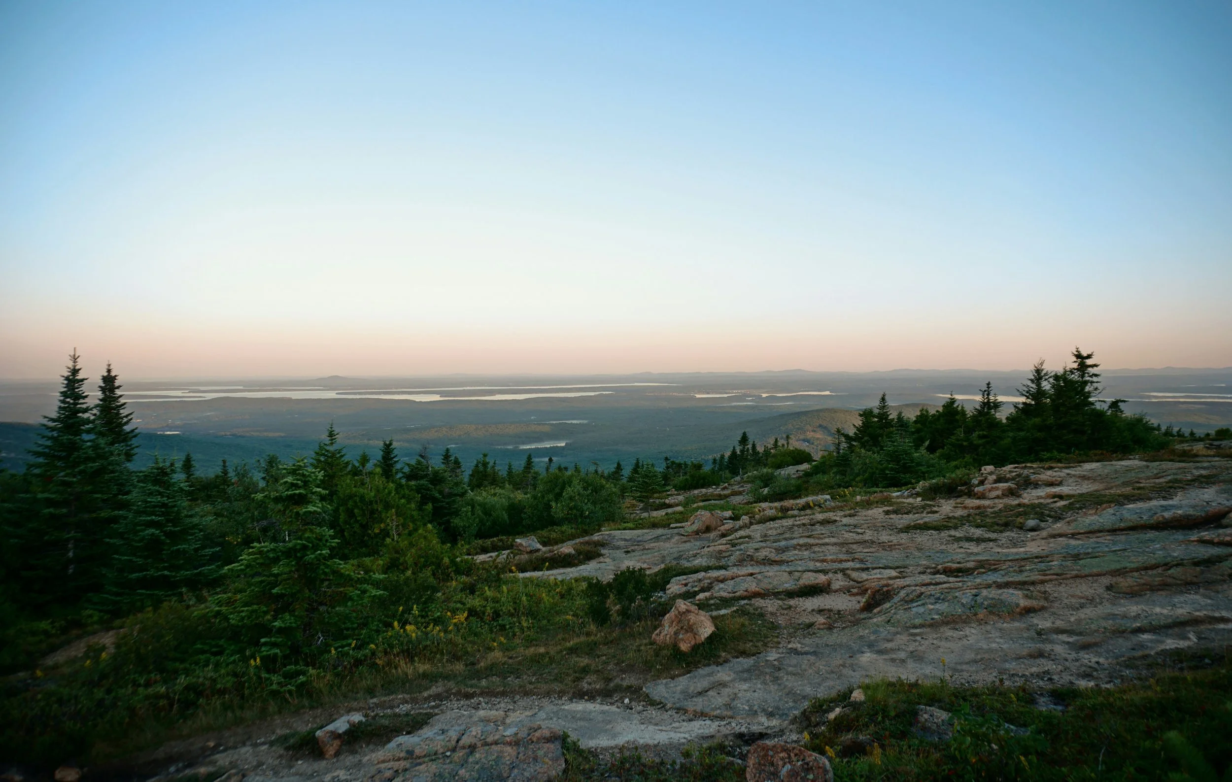 A landscape view of distant lakes and rolling hills seen from a rocky, forested hilltop with pine trees in the foreground during sunset or sunrise.