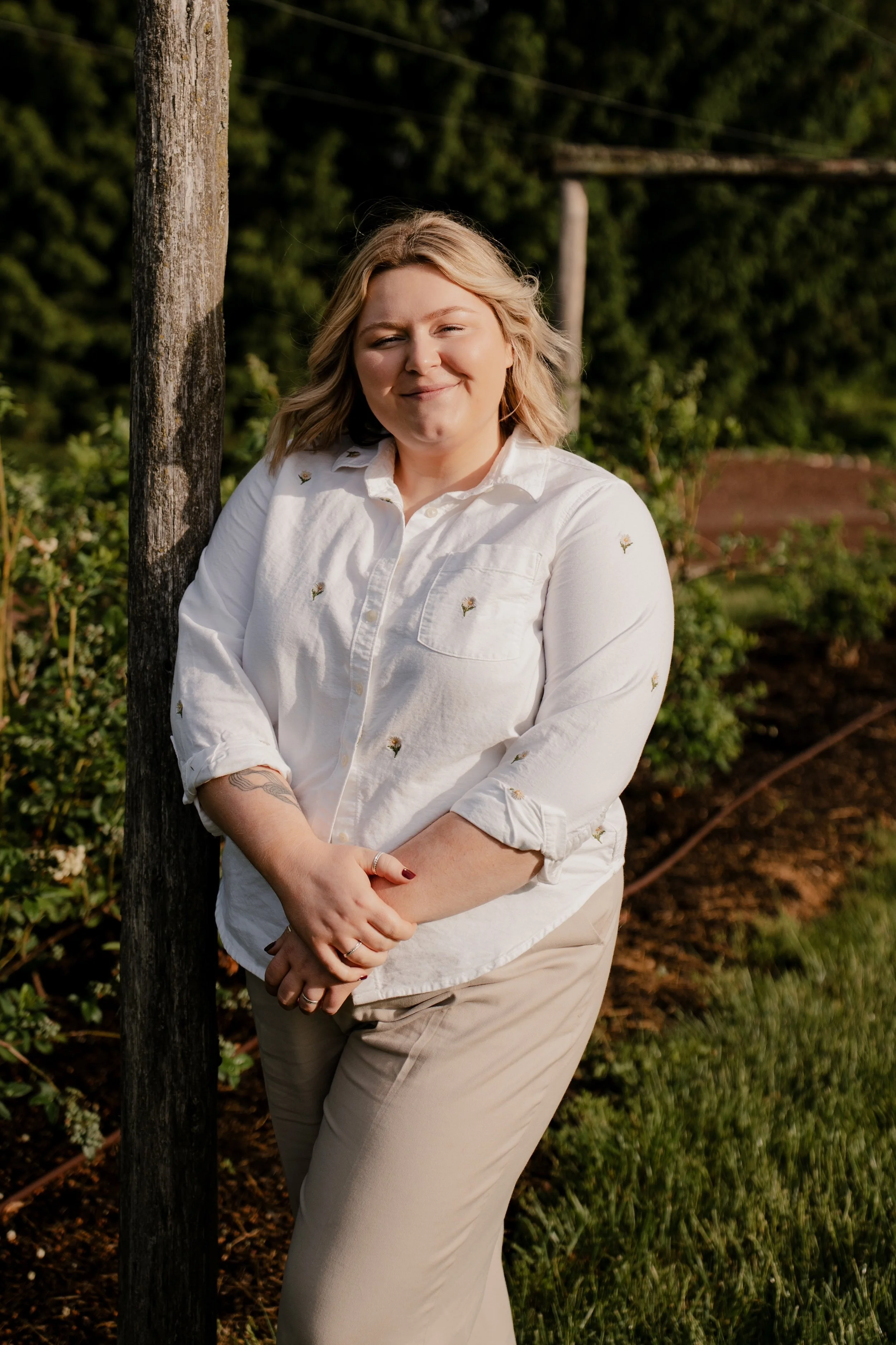 A woman with blonde hair, wearing a white shirt with small flower embroidery, leaning against a tree outdoors in a garden. She is smiling with eyes closed and has tattoos on her left arm.