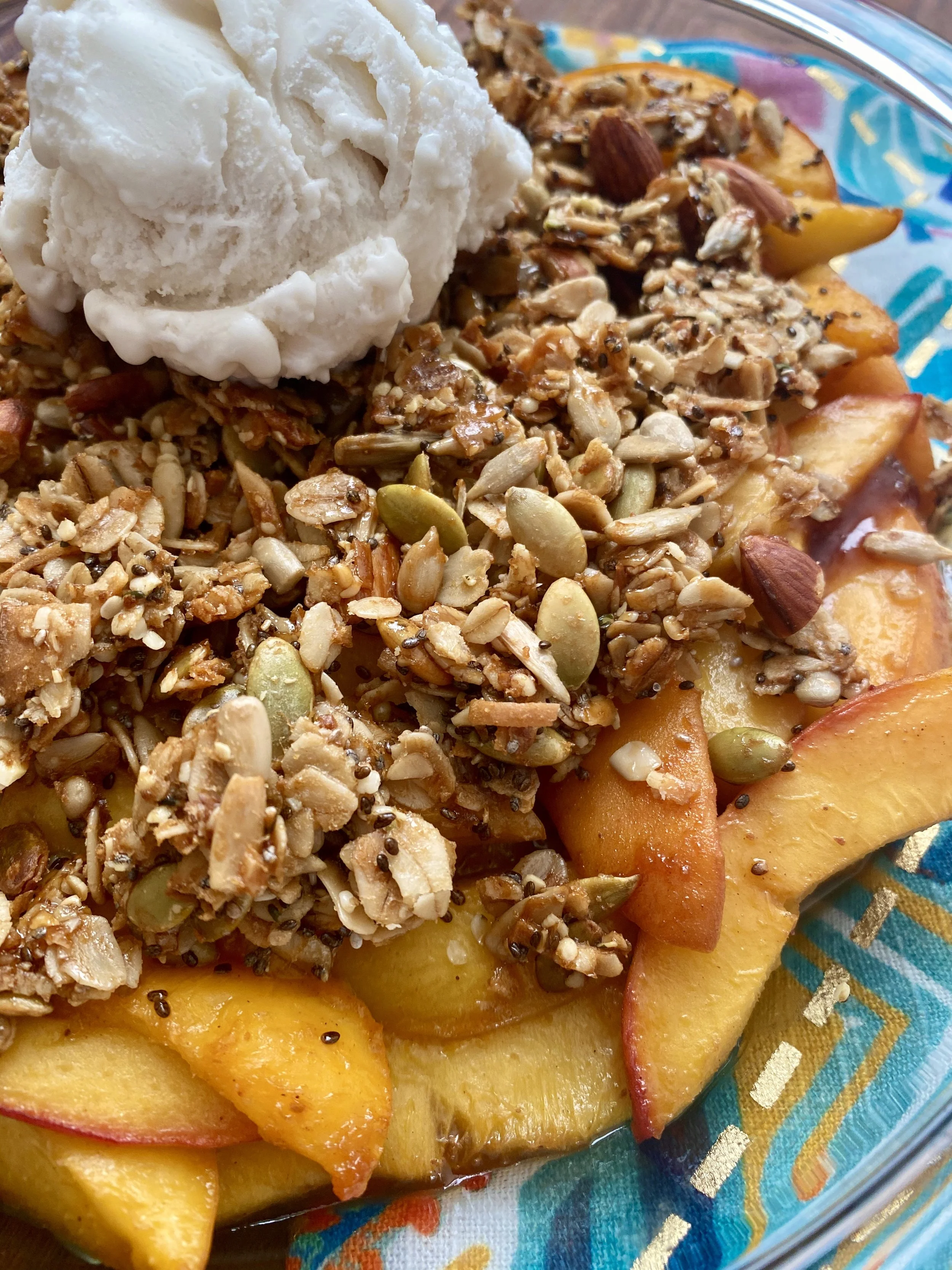 Baked sliced peaches topped with granola, pumpkin seeds, sunflower seeds, sliced almonds, and a scoop of vanilla ice cream in a glass dish.