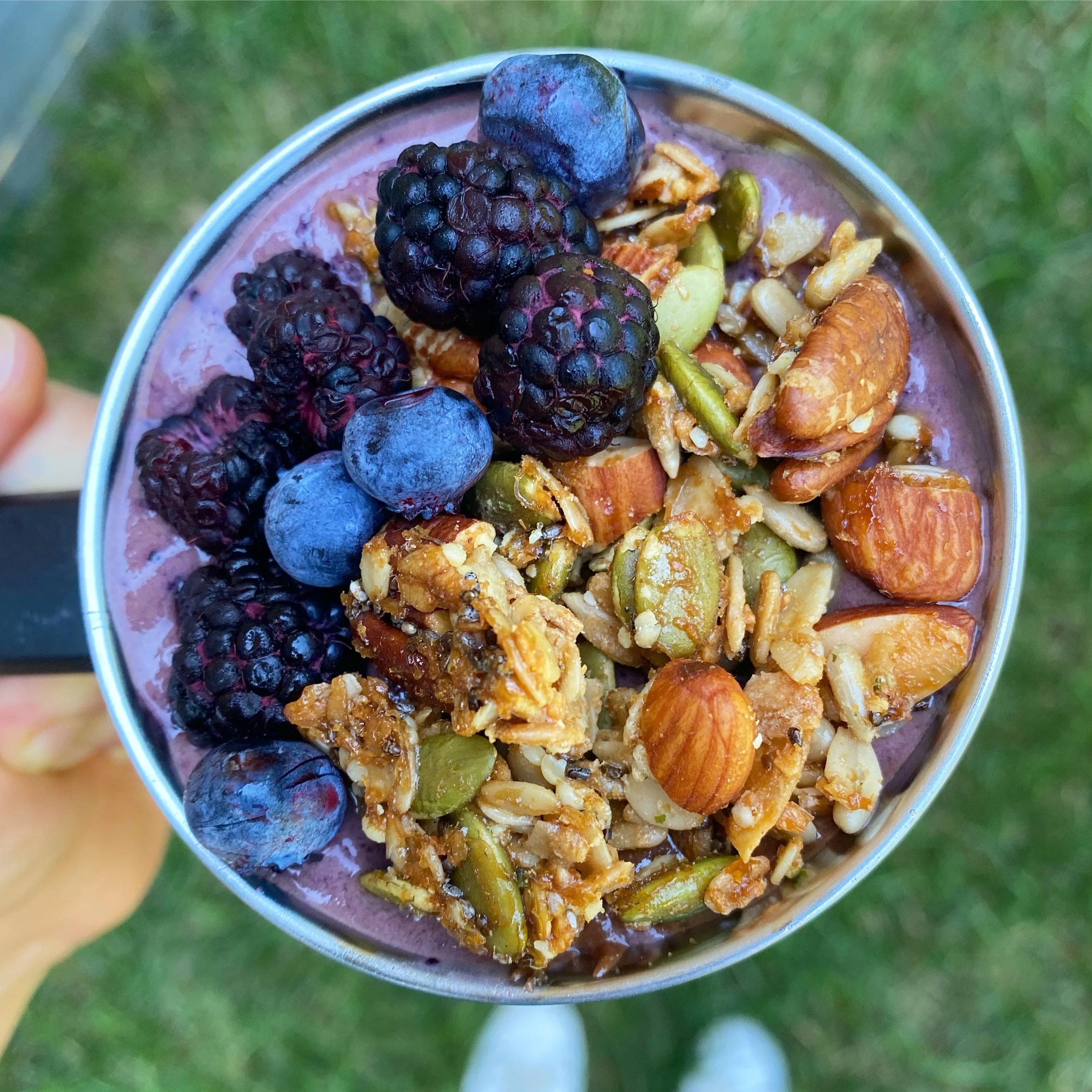 Close-up of a metal cup filled with granola, topped with blackberries and blueberries, with a green blurred background.