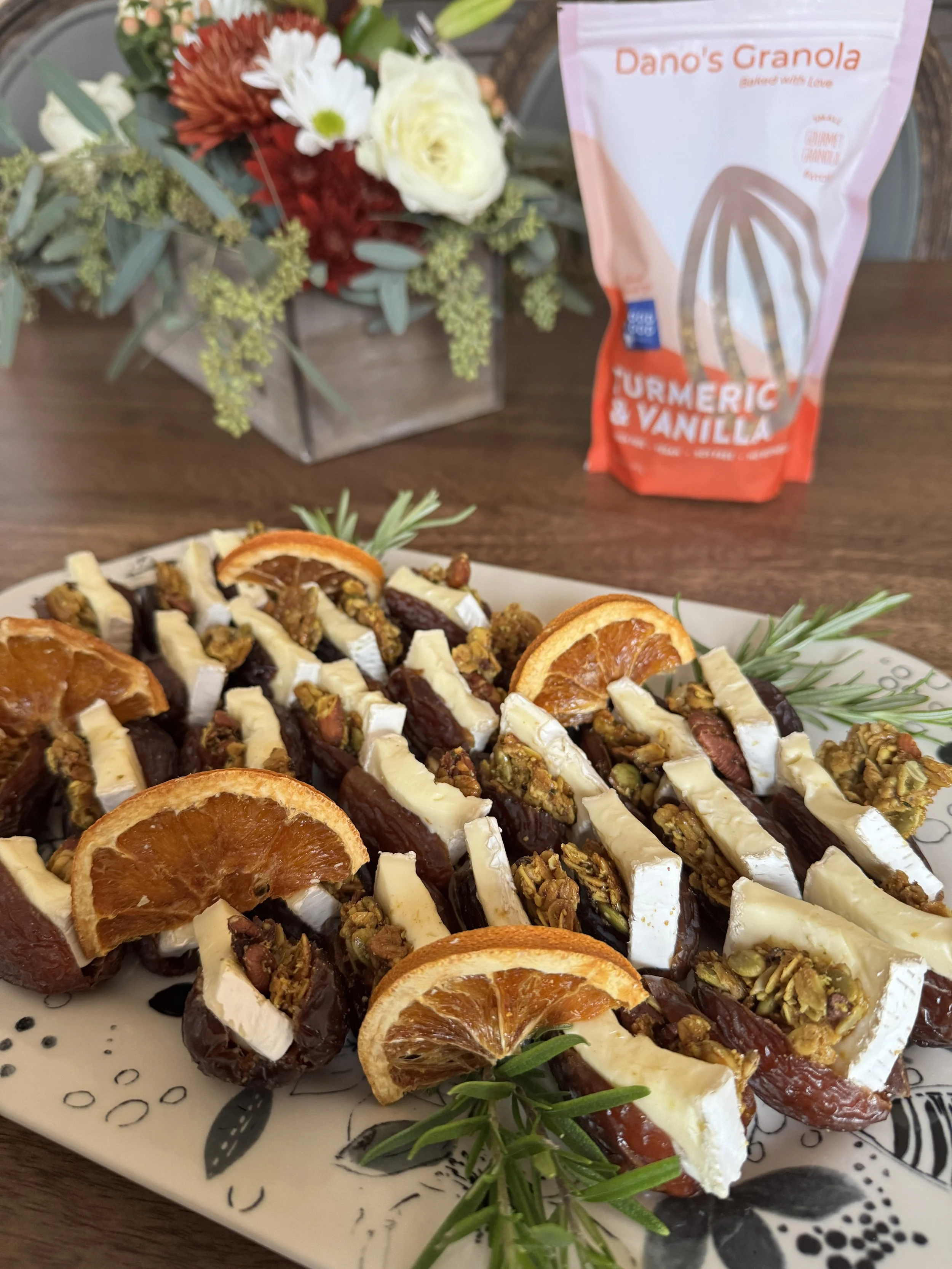 Plate of roasted vegetables topped with sliced dried oranges and garnished with rosemary, with a background of a bouquet of flowers and a bag of Dano's Granola with turmeric and vanilla flavor.