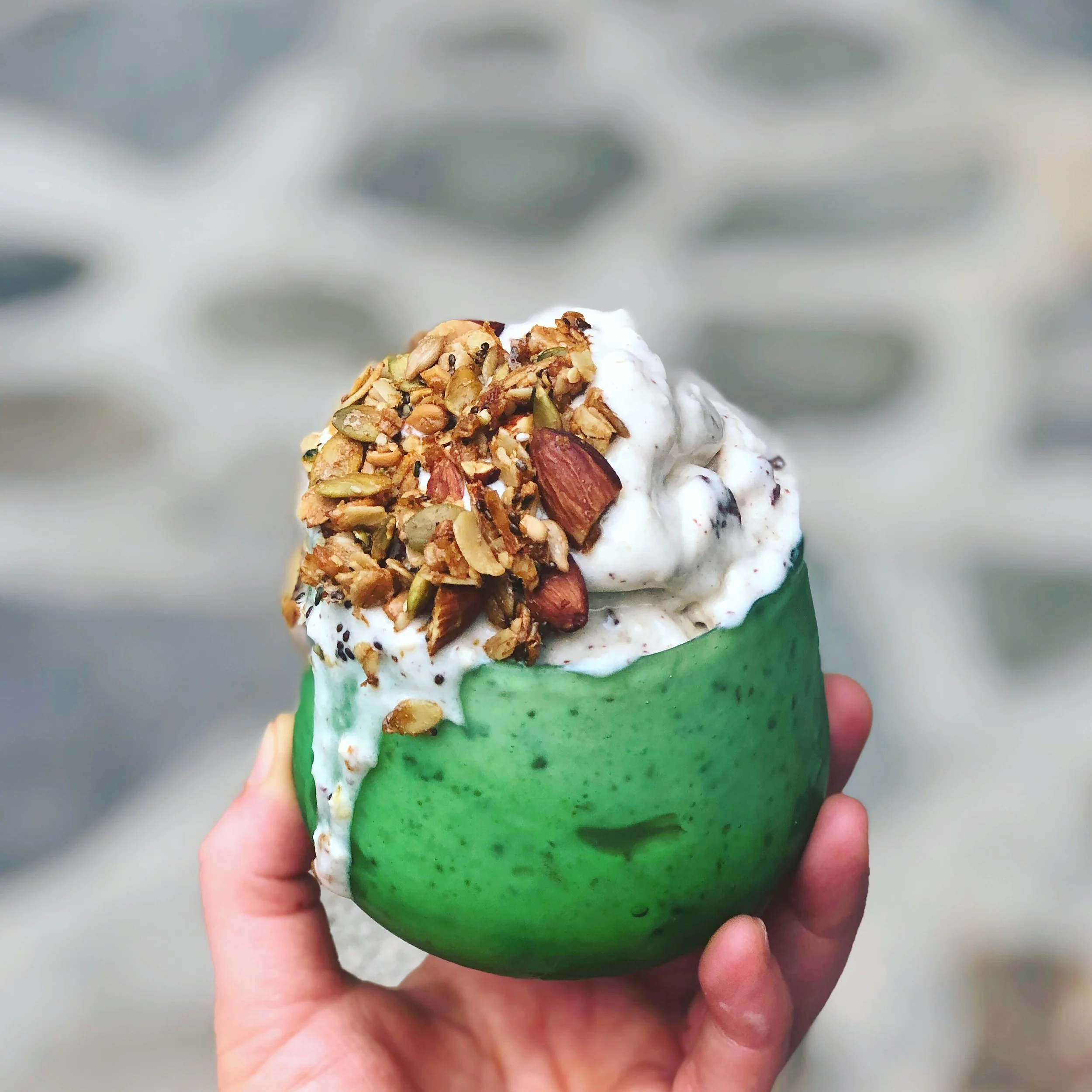 Close-up of a hand holding a green cup topped with yogurt and sprinkled with granola, nuts, and seeds, against a blurred background.