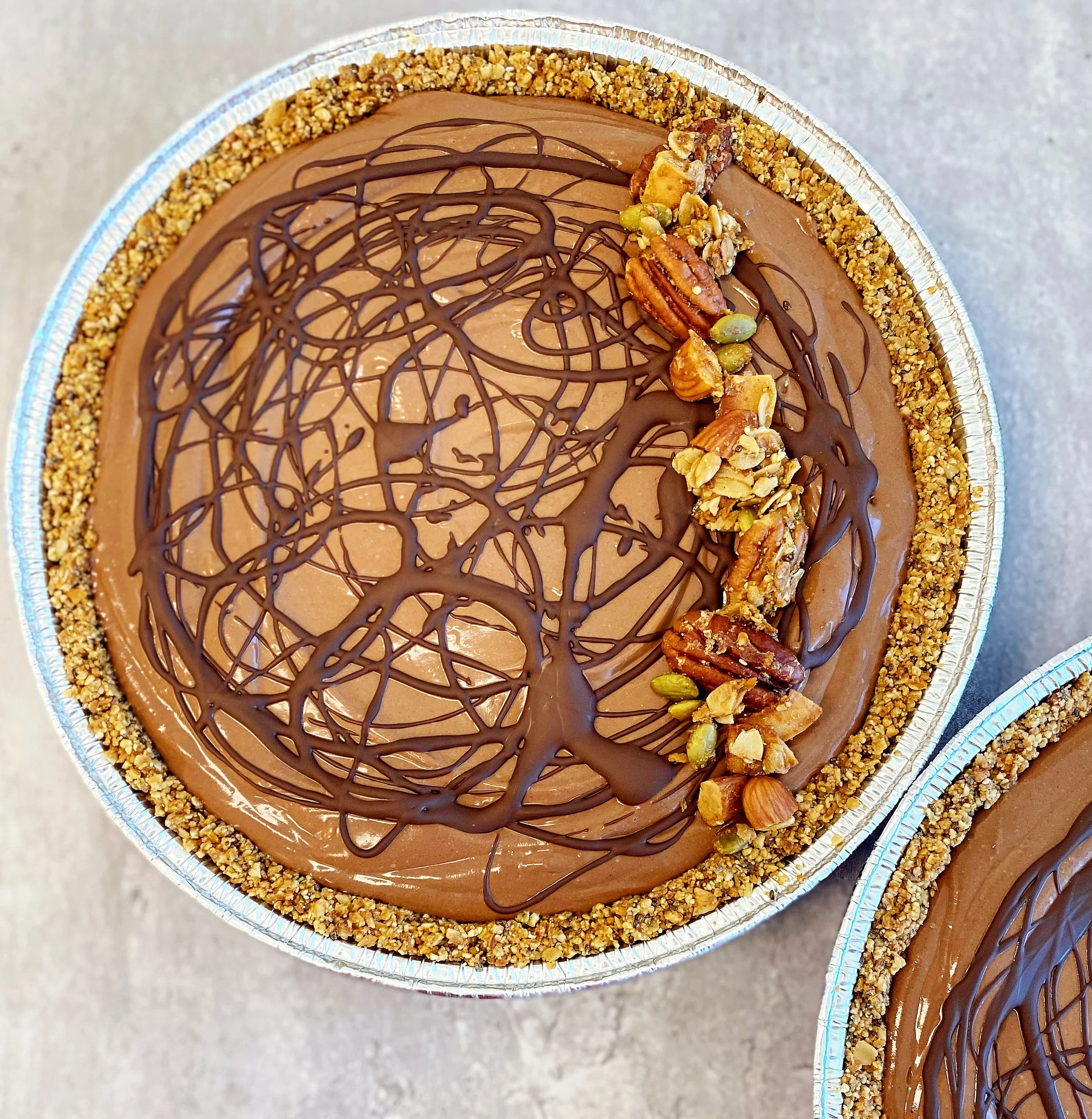 A round chocolate cake decorated with a drizzle of chocolate on top and surrounded by crushed nuts along the edge. It also has a row of mixed nuts and pistachios on one side.