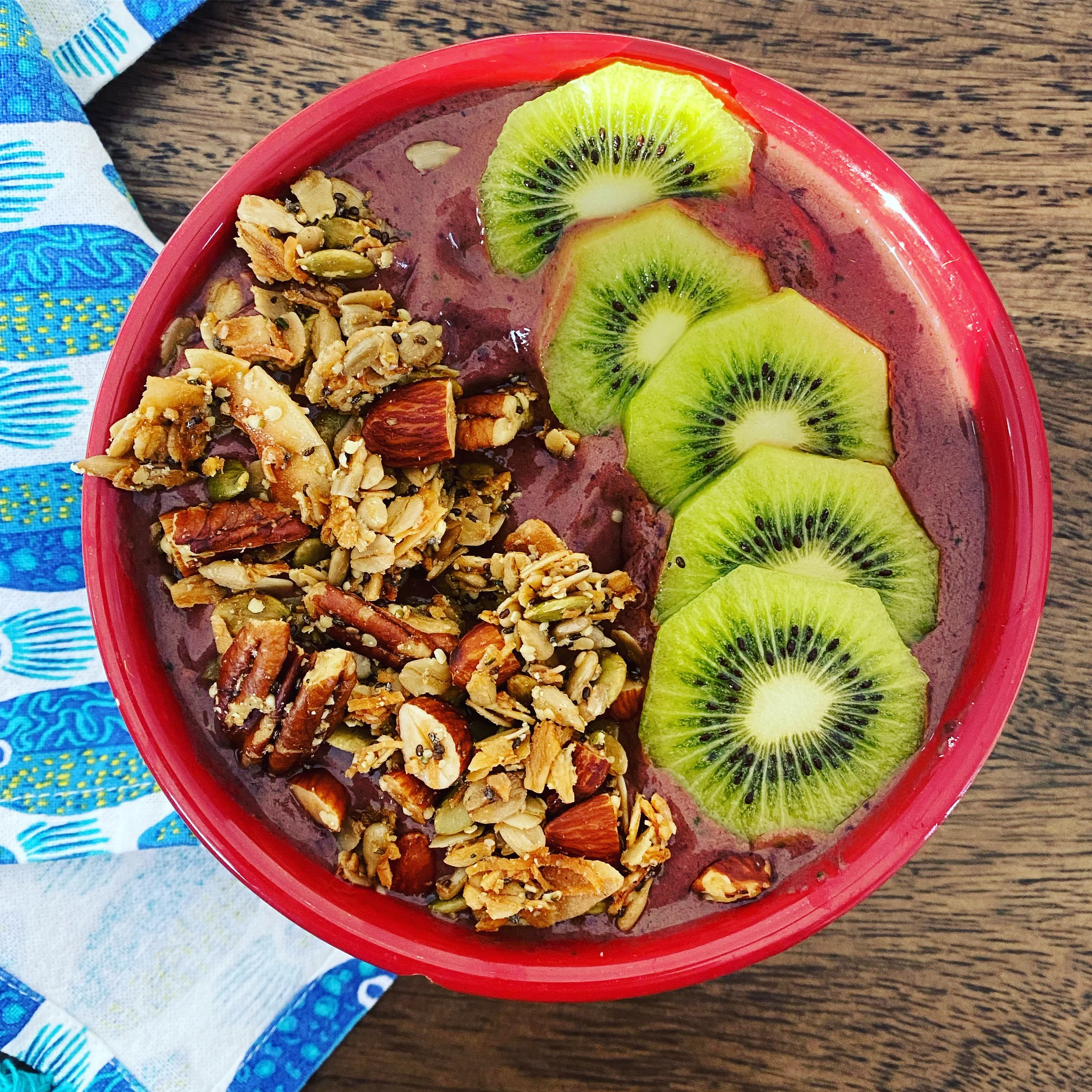 Close-up of a red bowl filled with assorted granola, topped with slices of kiwi fruit, on a wooden table with a colorful cloth nearby.