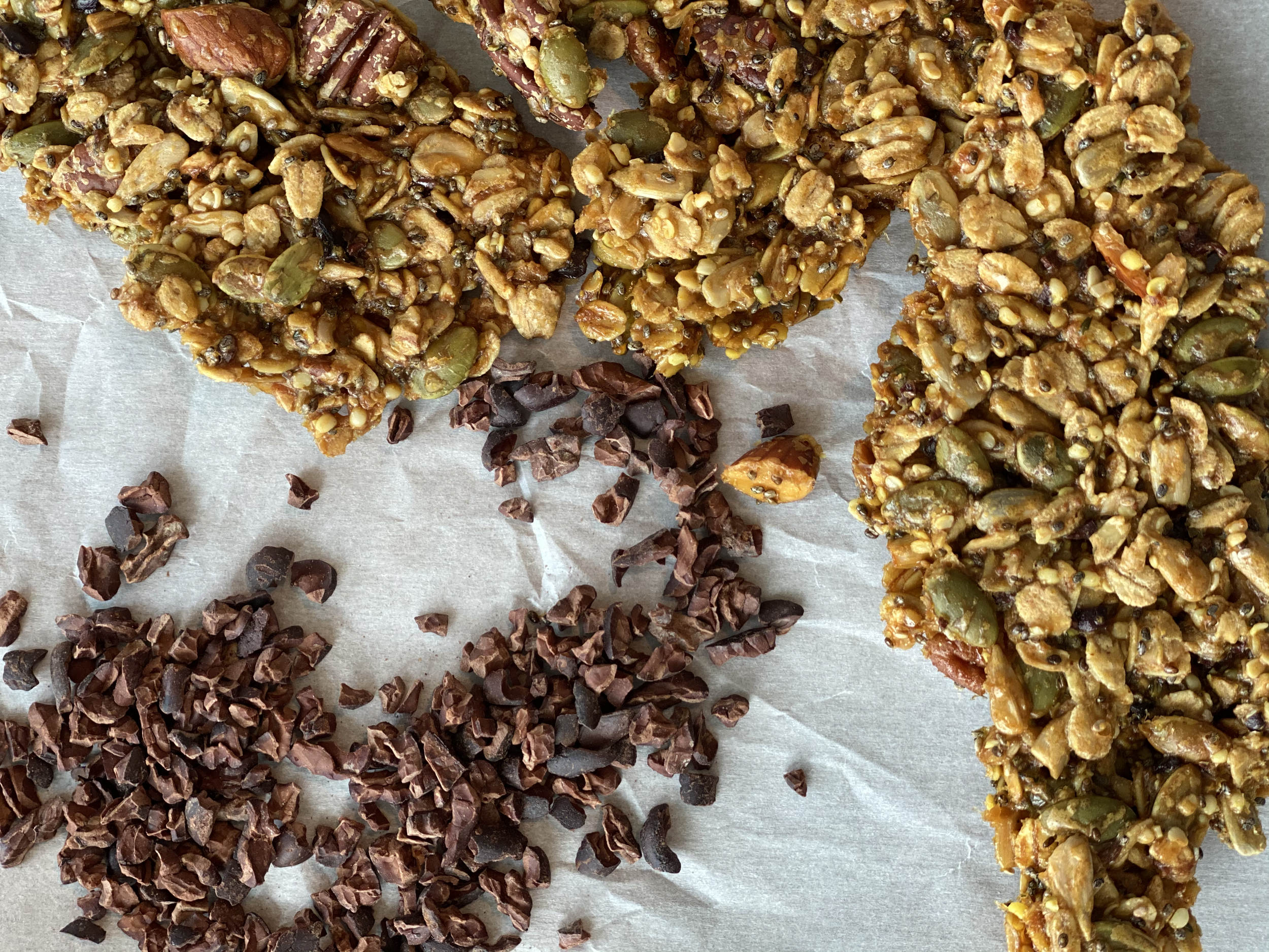 Close-up view of granola bars with pistachios, sunflower seeds, and dried fruit, along with small pieces of chopped chocolate on a parchment-lined surface.