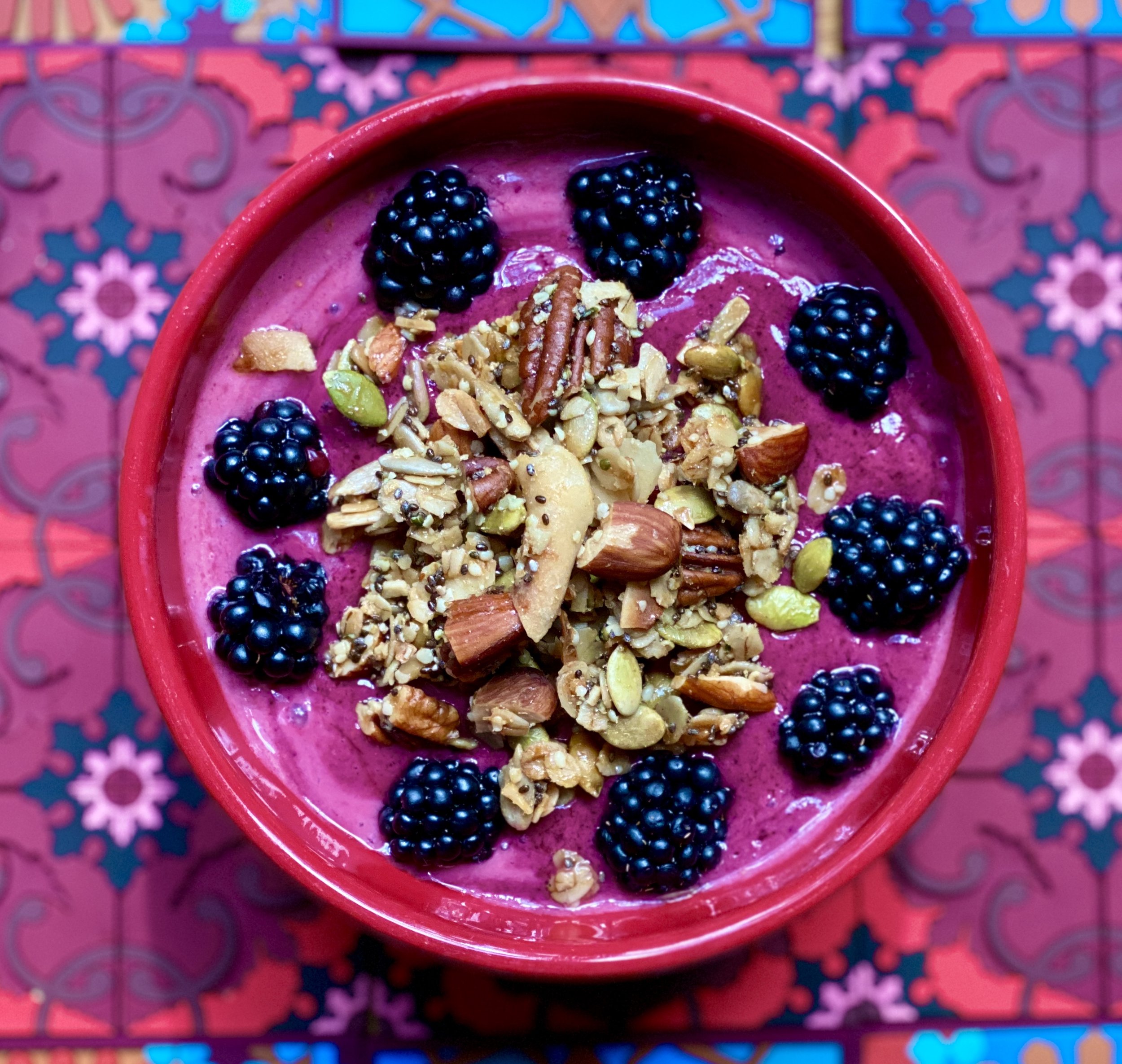 Close-up of a pink smoothie garnished with blackberries and mixed nuts in a red bowl, with a colorful, patterned background.