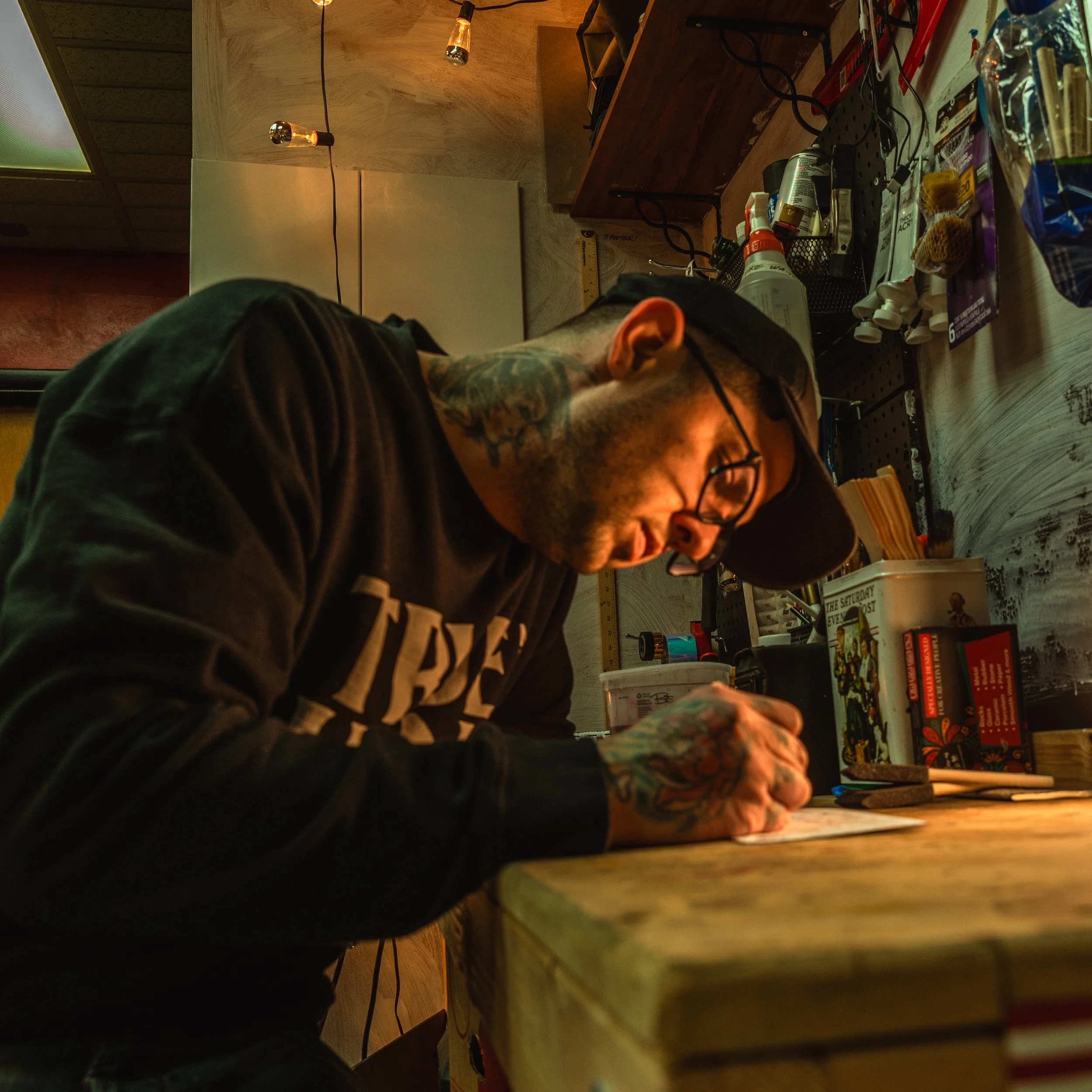 a tattooed man in a black hat and sweater wearing glasses sits at a desk surrounded by art supplies and warm lighting drawing
