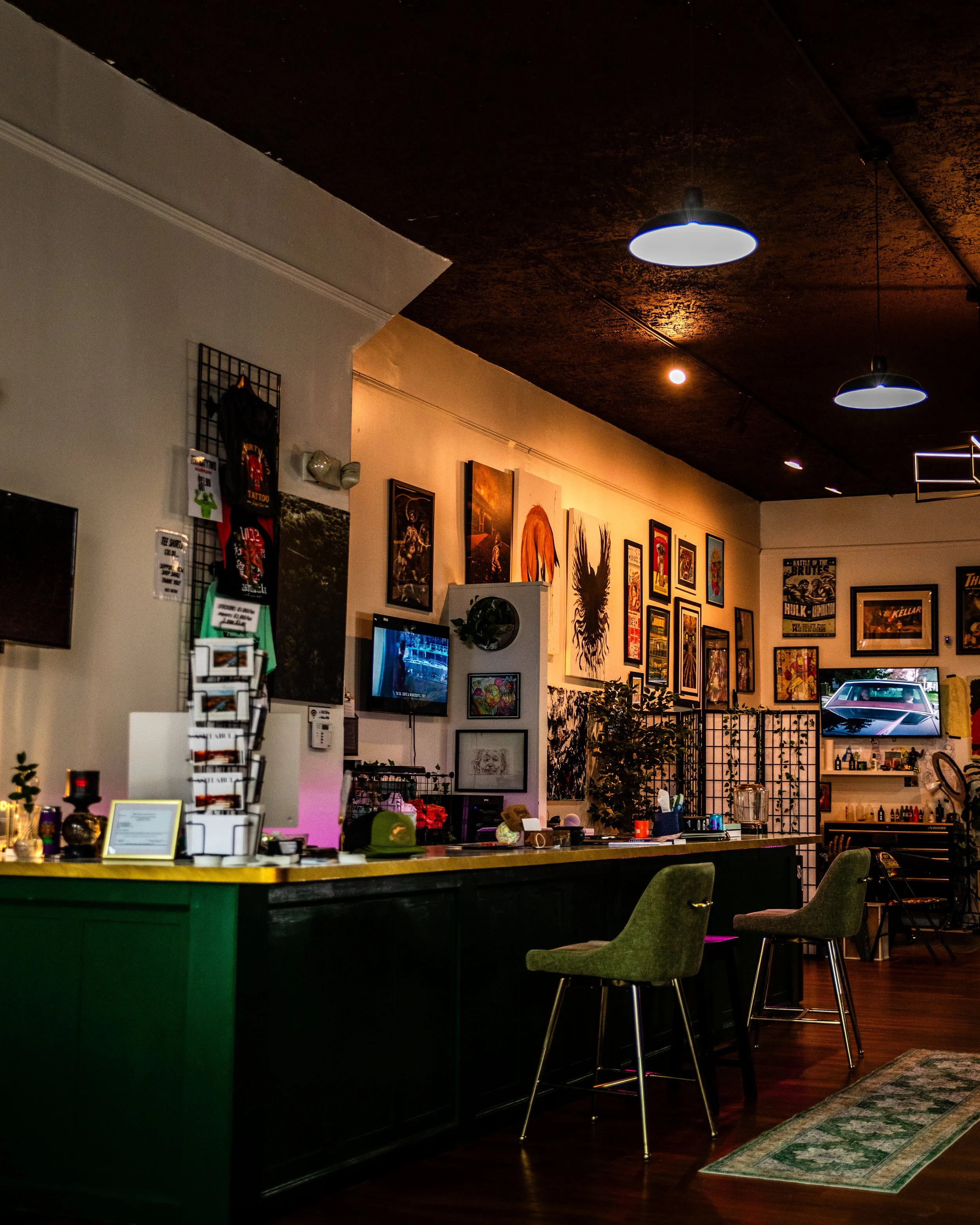 a view from the front waiting area of a charming and sophisticated tattoo shop with artwork displayed on the walls, televisions for clients entertainment and general realaxing atmosphere. greens and golds are found throughout creating a calm vibe.