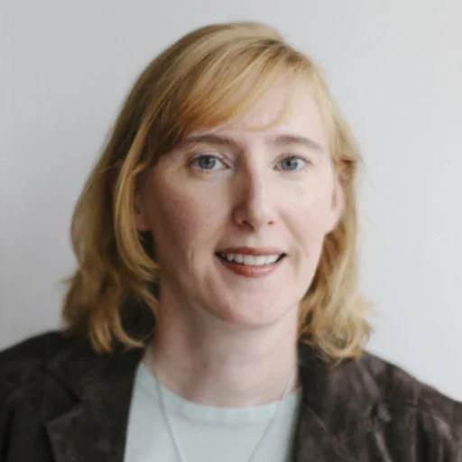 A headshot of Diana Gorman Jamrozik in front of grayish background. The person is a fair skinned and has a reddish blonde hair. The person is wearing a light blue top underneath what appears to be black jacket.