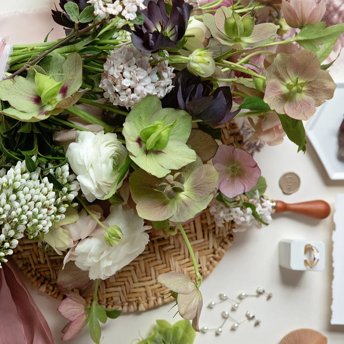 A bouquet of various flowers including white, pink, and dark purple blooms, arranged on a wicker mat on a white surface, with some jewelry and decorative items nearby.